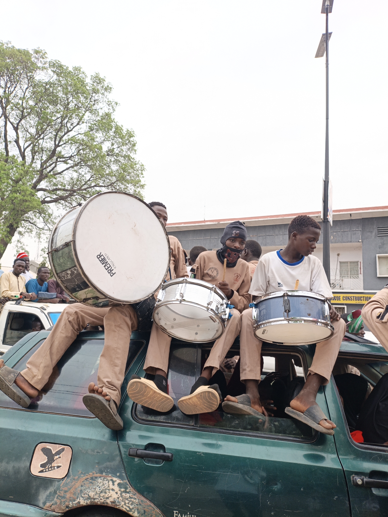 Four people are sitting on top of a dark-green, four-door car. Each person is wearing a tan-colored uniform. The person on the left is holding a large bass drum. The next person is holding a silver drum, and the person to the right of him is holding a blue drum. The person farthest to the right is partially out of view. Each person is wearing sandals. Other people are riding in the car with them. In the background are buildings and a streetlight. A tree is in the upper-left corner. A sticker is on the car with a bird of prey and the word "PEACE" written on it.