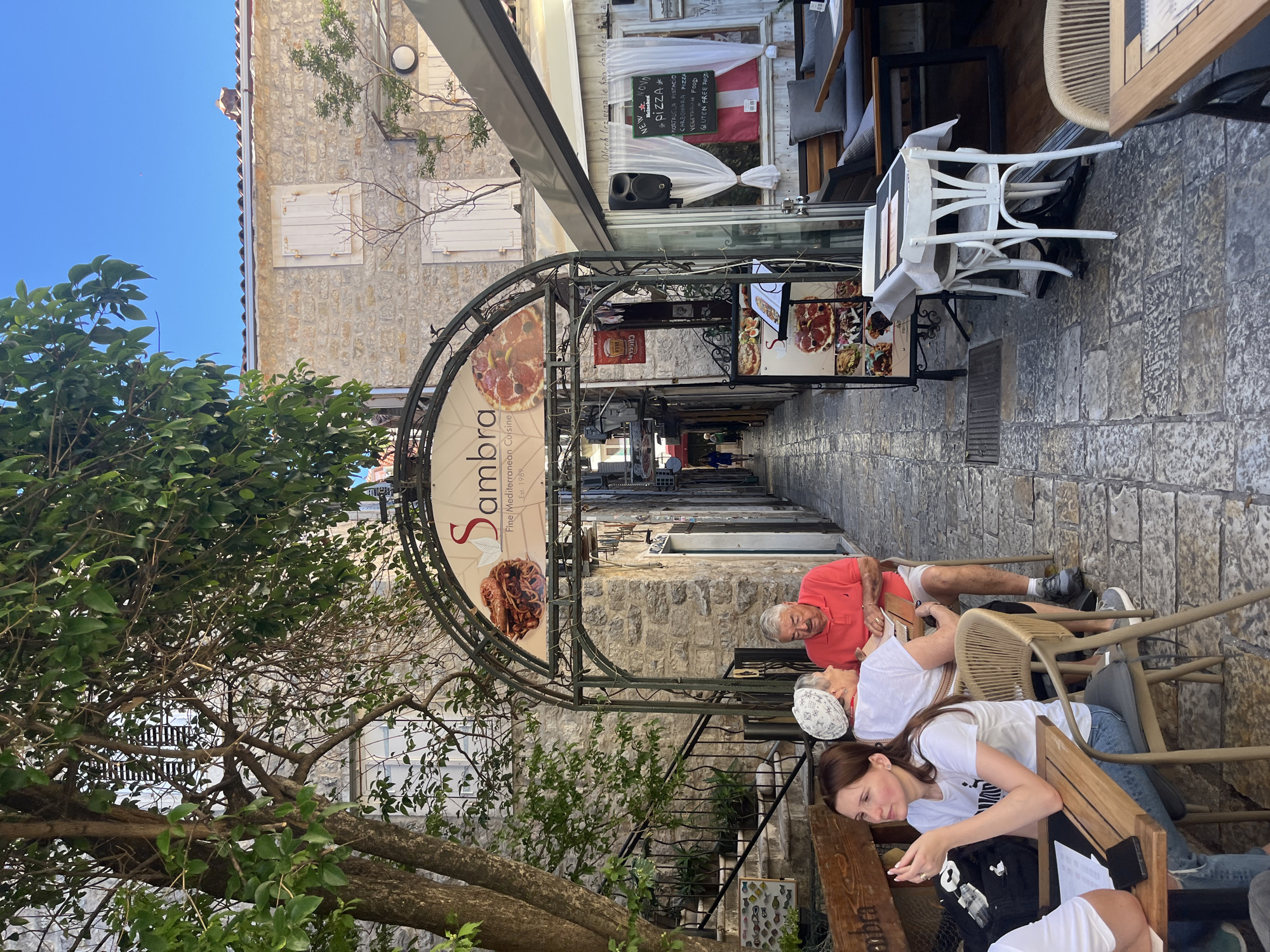 In Budva, Montenegro, a narrow cobblestone alleyway hosts an outdoor seating area for a restaurant. The restaurant's arched entrance features a sign with "Sambra Fine Mediterranean Cuisine Est. 1989" and images of food. In the foreground, a woman with brown hair, wearing a white t-shirt and ripped jeans, is seated at a table, holding a fork. Beside her, an older couple is also seated, with the man in a red polo shirt and the woman in a white t-shirt and headscarf, seemingly engaged in conversation or looking at a menu. The alleyway opens up to reveal more outdoor seating and a glimpse of people walking further down the lane. The buildings flanking the alley are constructed of stone, and lush green trees provide shade. The overall scene depicts a relaxed, sun-drenched atmosphere.