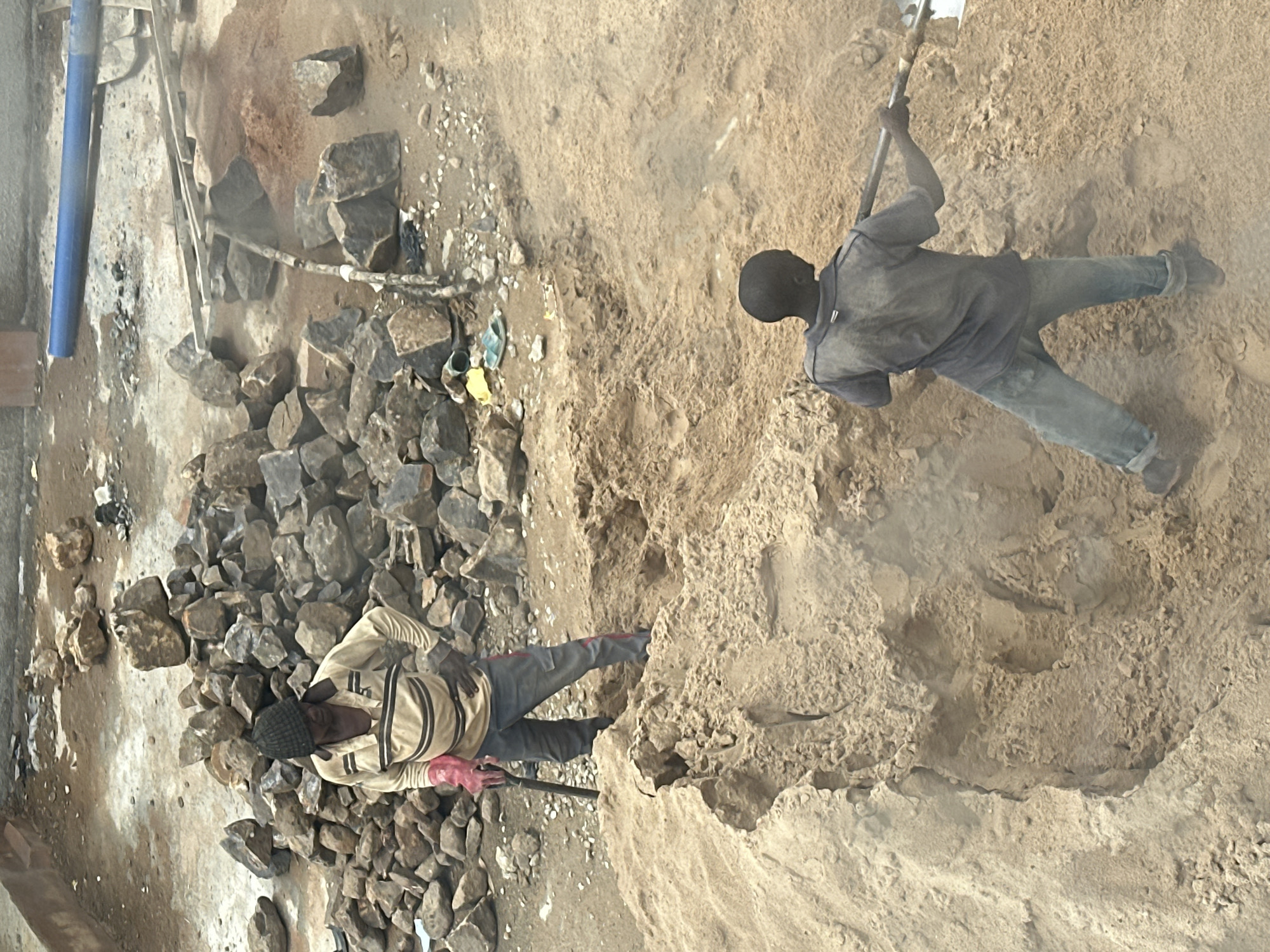 In Barakin Lerawi, Nigeria, two individuals are engaged in manual labor involving sand. The scene is outdoors, with a dusty, rough terrain characterized by piles of sand and large rocks.
In the foreground, a person wearing a dark blue shirt and denim jeans is actively shoveling sand, creating a dust cloud. The individual is holding a shovel with both hands, positioned to move the sand.
In the mid-ground, another person, dressed in a tan long-sleeved shirt with horizontal dark stripes, dark pants with red accents, and a dark beanie, stands amidst a pile of sand and rocks. This individual is holding a shovel vertically with their left hand. Red gloves are visible on their hands. They appear to be observing or resting.
The environment includes a significant amount of loose sand and scattered rocks of various sizes. In the background, a blue pipe is visible, along with more rocks and debris, suggesting a construction or landscaping site. The overall atmosphere is arid and industrial.