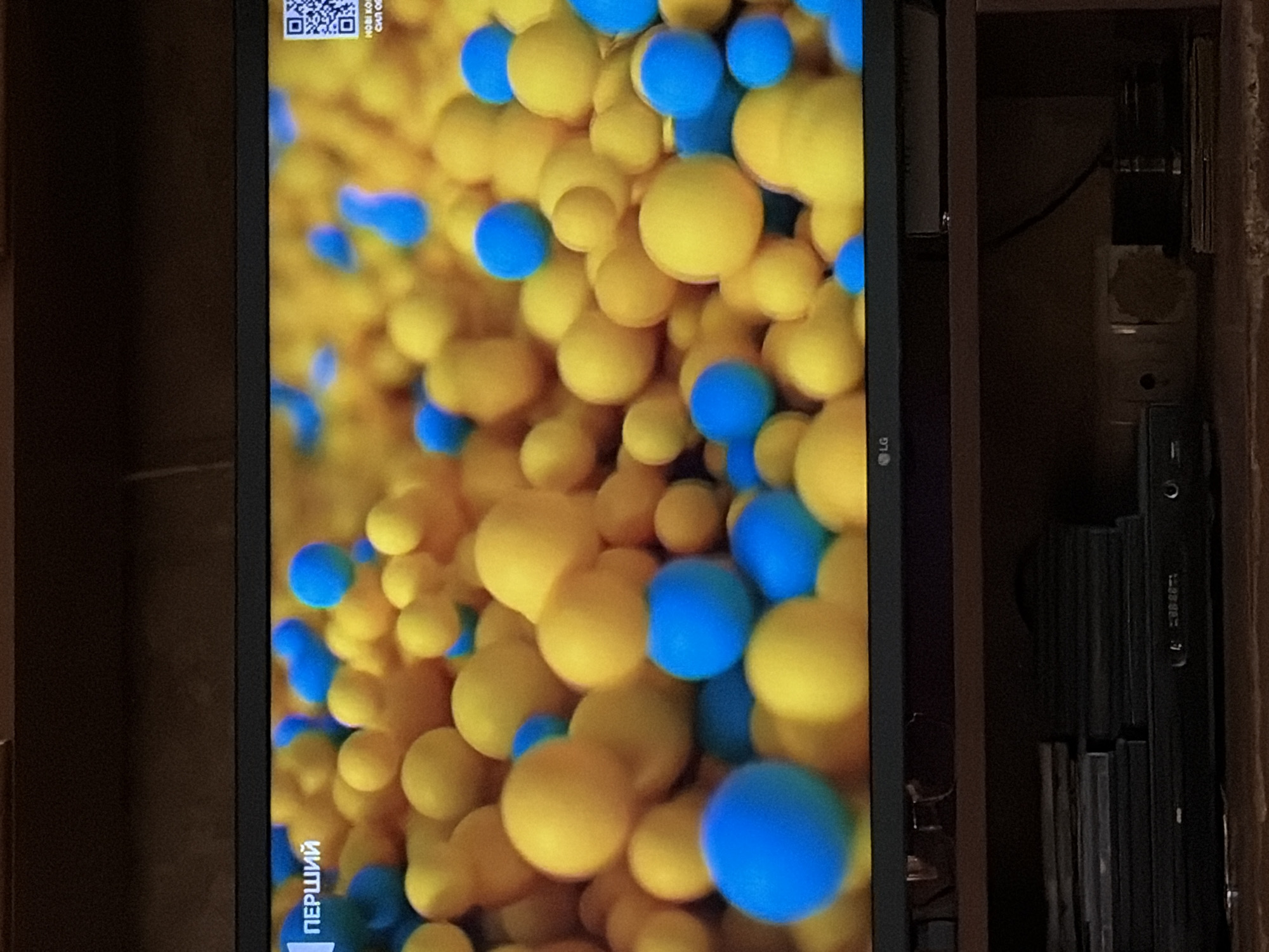 The image shows a close-up of a television screen displaying a colorful animation of yellow and blue spheres. Text in Cyrillic, reading "ПЕРШИЙ," is visible in the upper left corner of the screen. In the upper right corner, a QR code is displayed. The television is set on a dark shelf, beneath which are stacks of black discs, likely CDs or DVDs, and a black electronic device. The overall environment suggests an indoor setting, likely a living room. No people are visible in the image.