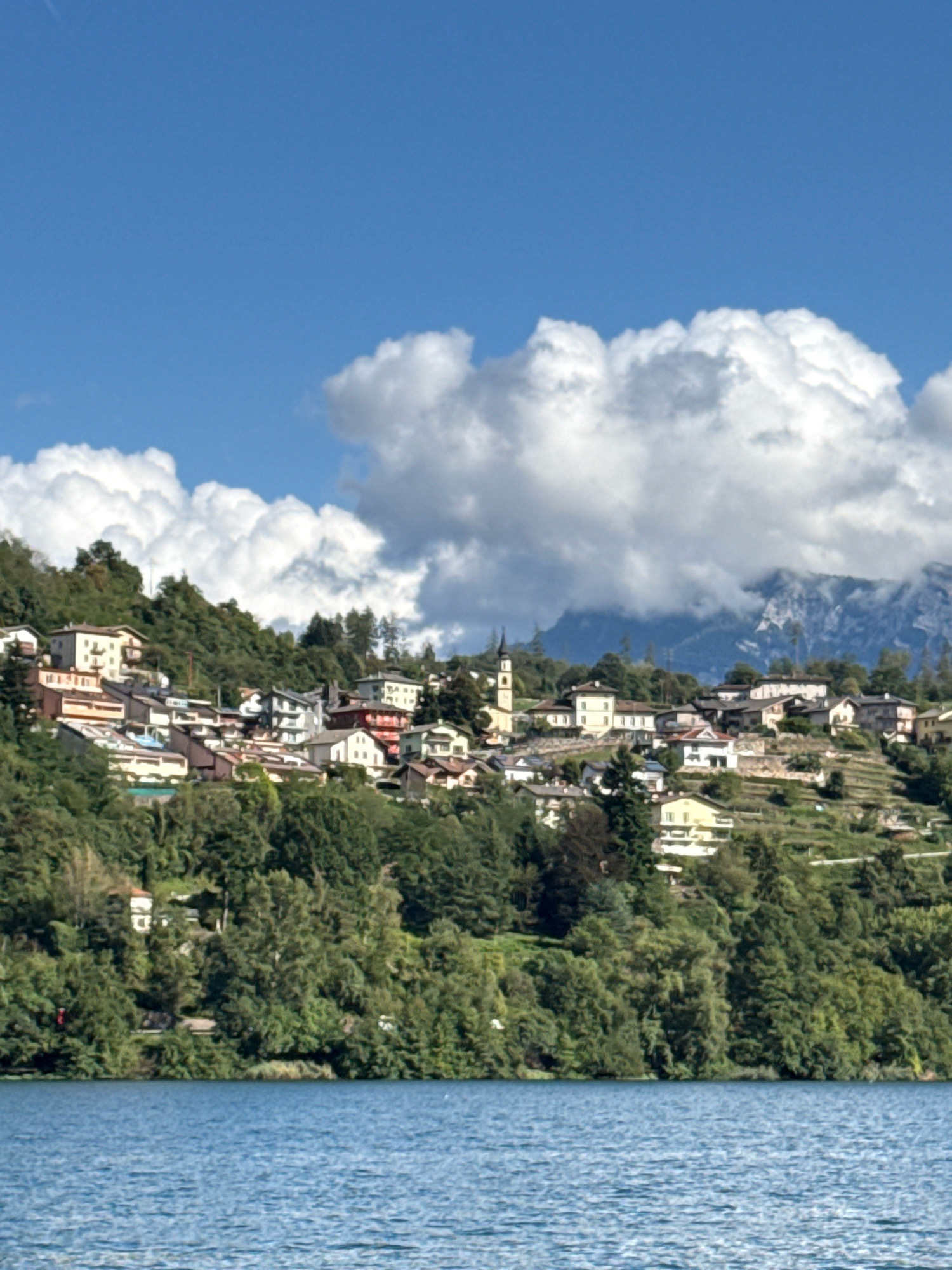 A wide-angle, eye-level shot captures a picturesque view of a lakeside town nestled on a verdant hillside under a bright blue sky dotted with fluffy white clouds. The water in the foreground is a vibrant blue with gentle ripples, reflecting the sunny day. The hillside is densely covered with lush green trees, interspersed with terraced vineyards and a charming cluster of residential buildings. The architecture is varied, featuring multi-story homes with colorful facades, terracotta roofs, and balconies. A prominent church spire with a dark roof rises above the buildings, adding a classic touch to the village. In the background, misty mountains loom under the dramatic clouds, hinting at the dramatic natural landscape of the Trentino region. The scene is tranquil and idyllic, showcasing the beauty of a small Italian town in harmony with its natural surroundings. Based on the context of Pergine Valsugana, Italy, this view likely depicts a scene along Lake Caldonazzo or Lake Levico, popular tourist destinations in the Trentino region.