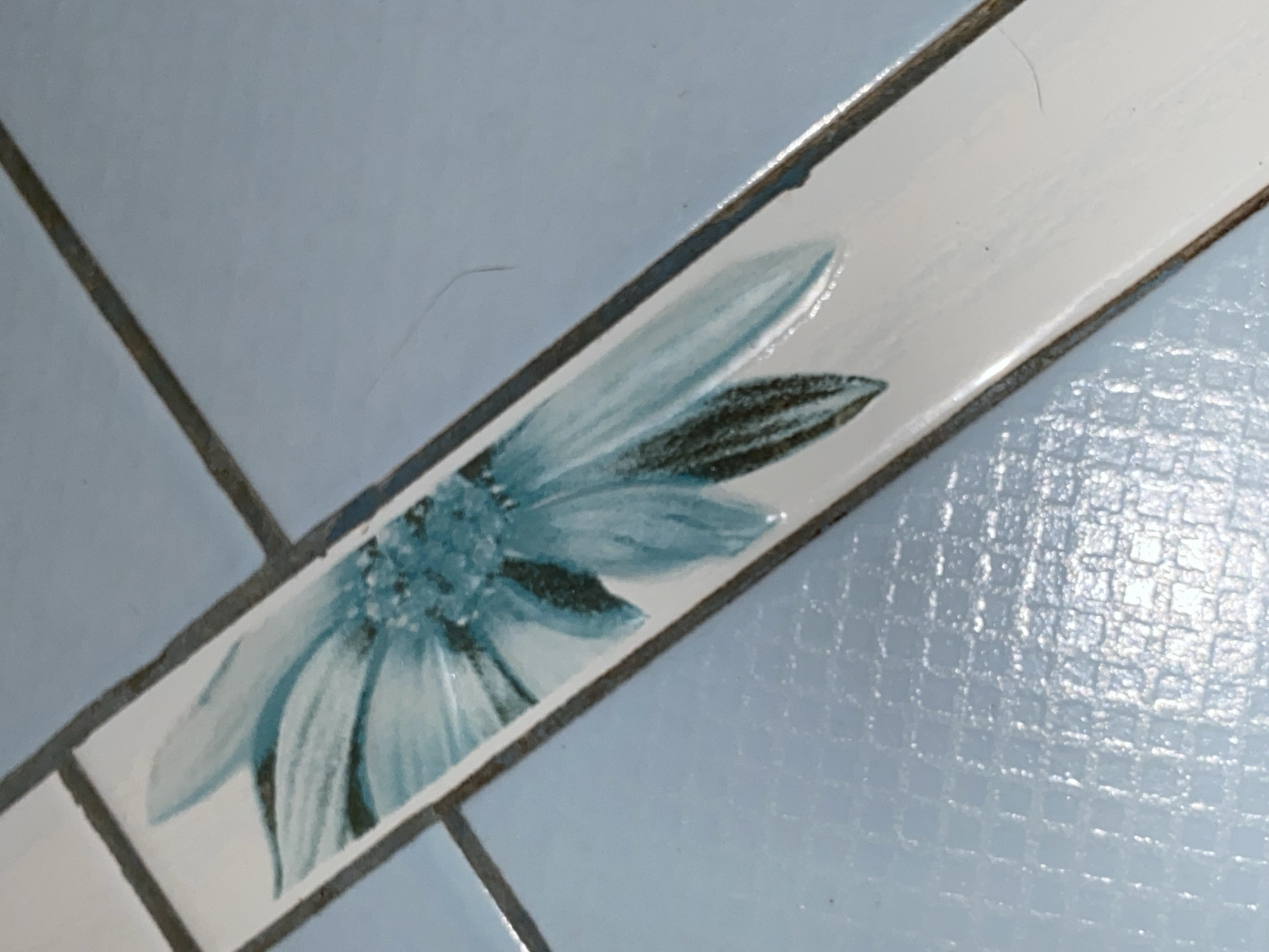 The image shows a close-up of ceramic tiles. Some tiles are light blue, others are white with a painted blue flower design. The tiles are arranged in a grid pattern with visible grout lines. The texture of the blue tiles appears slightly textured or patterned. The flower design is positioned on the white tile. It features petals of different shades of blue. There is no indication in the image that shows this photo to be from the city of Chaikovsky, Russia.