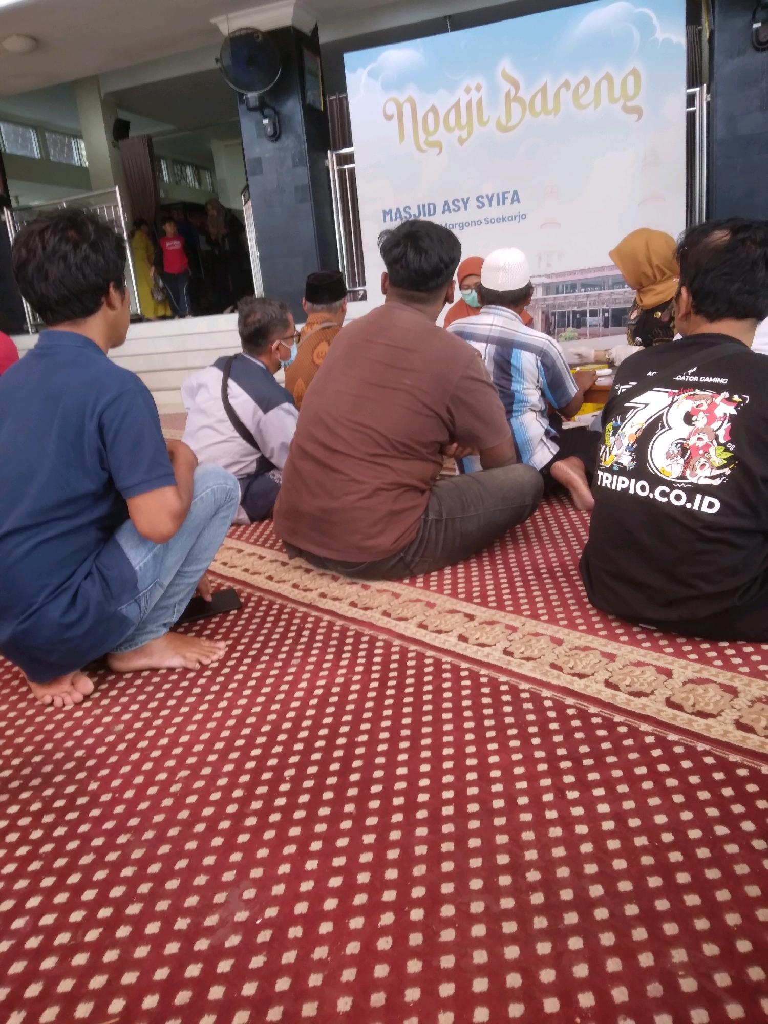 The image captures an indoor scene, likely a community gathering at a mosque in Desa Pamijen, Kecamatan Sokaraja, Indonesia. A large banner with the text "Ngaji Bareng" and "MASJID ASY SYIFA Margono Soekarjo" is prominently displayed. Several individuals are seated on a red carpet with a white patterned design, facing away from the camera.
In the foreground on the left, a person wearing a blue t-shirt and jeans is squatting, barefoot, with a black object, possibly a phone, next to them. To their right, another individual in a brown t-shirt and dark pants is also seated. Further back, several people are visible, some wearing traditional religious headwear. One person in the mid-ground is wearing a striped blue and white shirt and a white cap. On the right side of the image, a person in a black t-shirt with a large graphic and text, including "TRIPLO.CO.ID," is seated.
The environment appears to be the prayer hall of a mosque, indicated by the carpet and the presence of a banner for a religious event. The lighting suggests it is daytime. The overall atmosphere seems to be one of a communal religious activity.