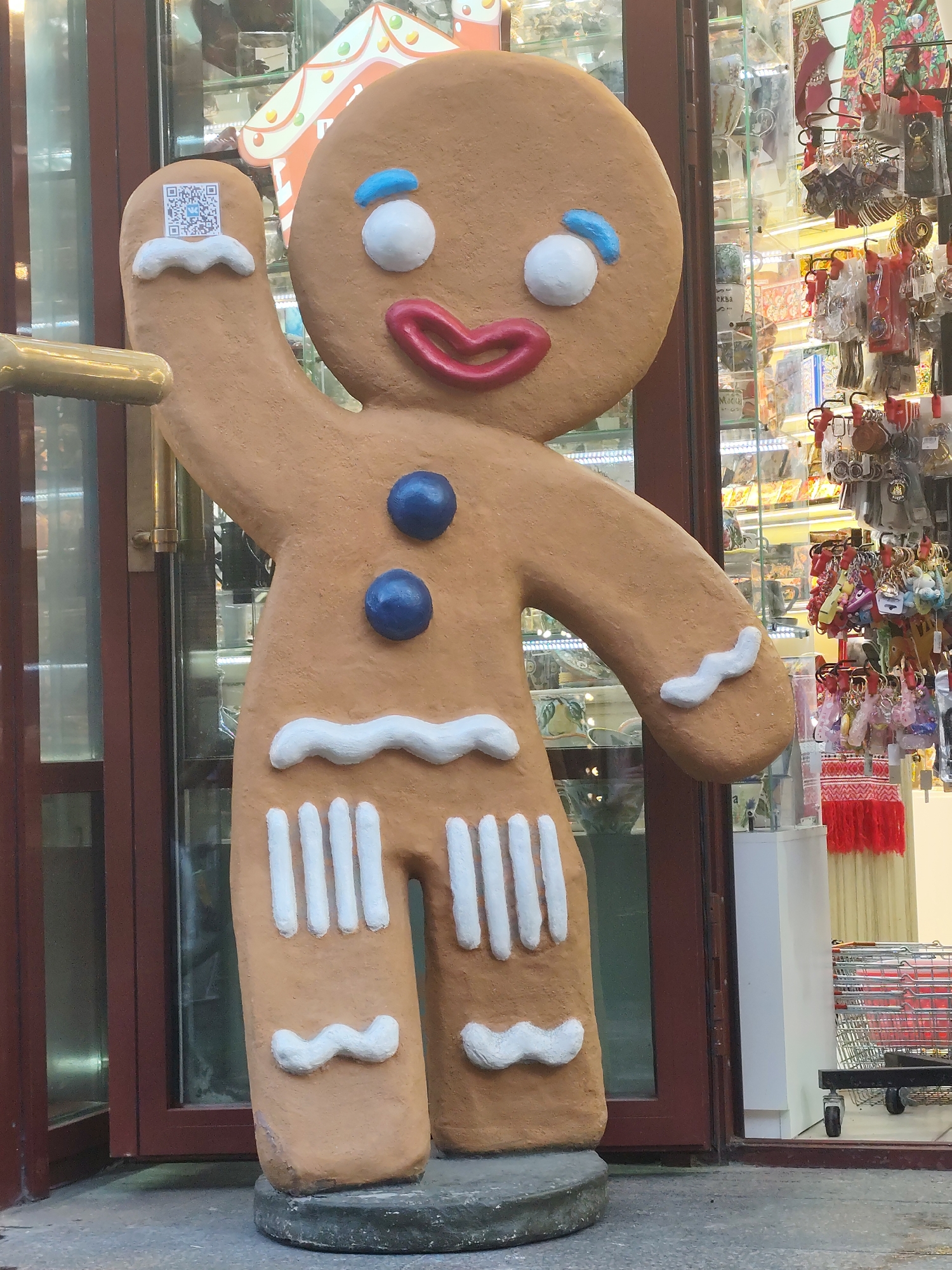 A large gingerbread man statue stands in front of a storefront in Moscow, Russia. The statue is brown with white icing trim and has blue eyes and buttons. A QR code is affixed to its raised hand. The storefront behind the statue is filled with merchandise. A red shopping cart is visible through the glass. The building has a maroon-painted frame. The statue is on a grey cement base. The sidewalk is grey.