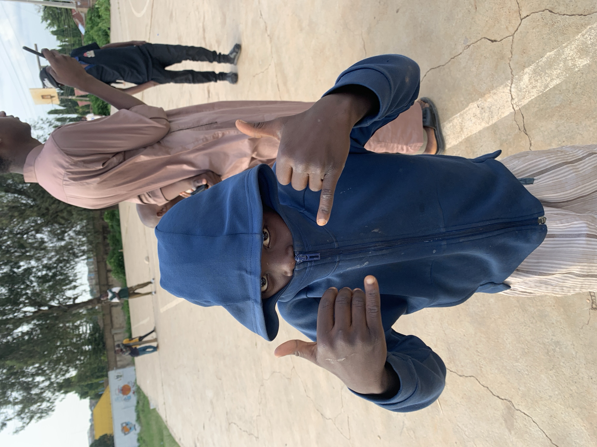 In Jos, Nigeria, a young person wearing a blue hooded jacket and striped pants stands on a cracked concrete court, giving a "shaka" hand gesture with both hands. Their hood is pulled up, partially obscuring their face, with only their eyes visible. In the background, other individuals are present, some standing and others possibly walking. One person in a pink, long-sleeved garment holds a phone, and another in a black shirt and pants stands with hands in their pockets. A basketball hoop is visible in the distance, along with trees, buildings, and a wall with colorful drawings. The lighting suggests it is daytime, possibly late afternoon.