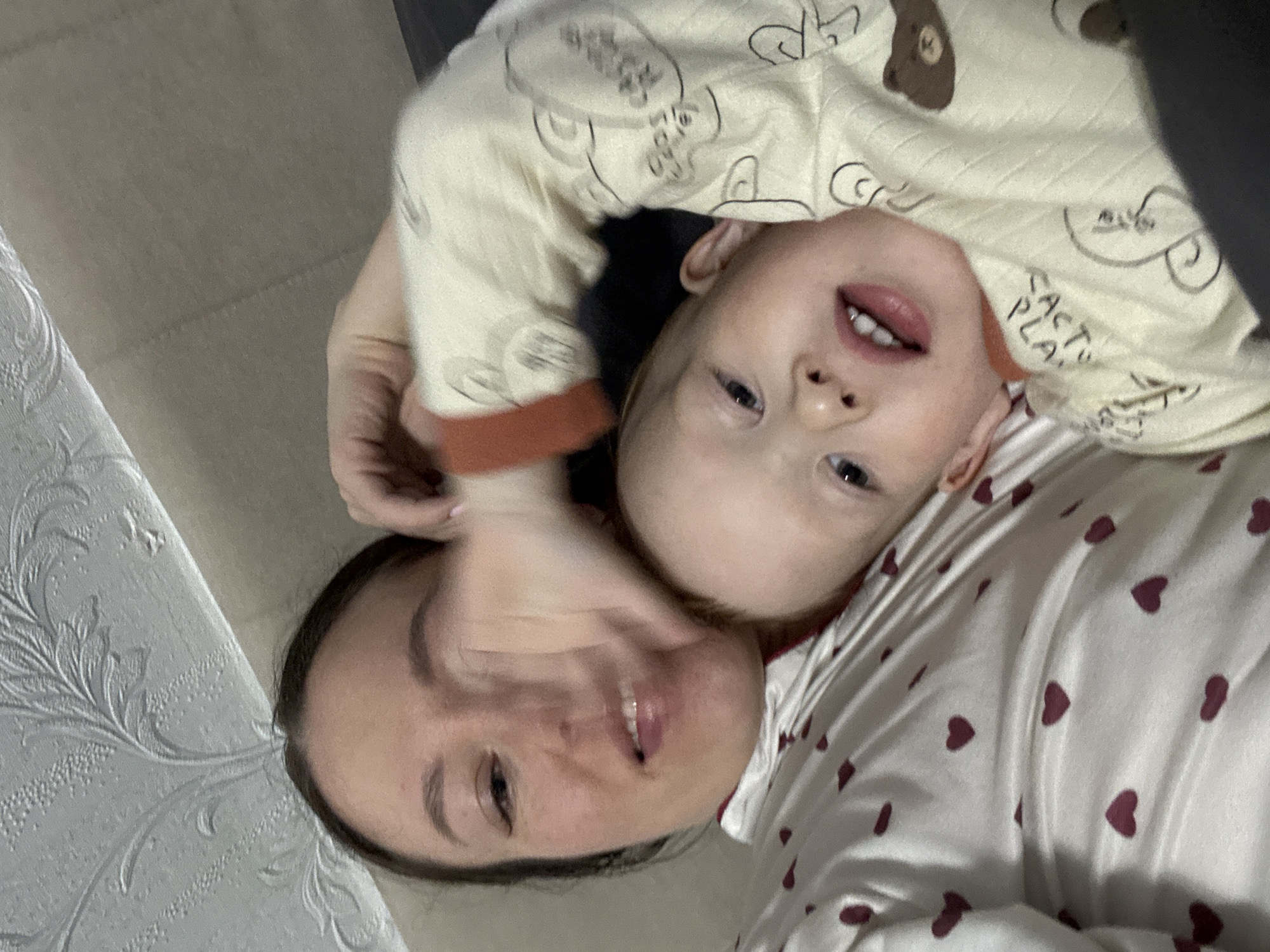 A selfie shows a young child and adult laying on their backs, presumably on a bed or soft surface, in Ферма, Russia. The child, who appears to be a toddler, is wearing pajamas with cartoon-like images, possibly bears. The adult is wearing pajamas with red hearts on them. Both are smiling slightly at the camera. The adult appears to have brown hair pulled back, and their hand is raised above the child's head, possibly supporting their position. The background is blurry but shows parts of a wall with embossed texture and a flat surface.