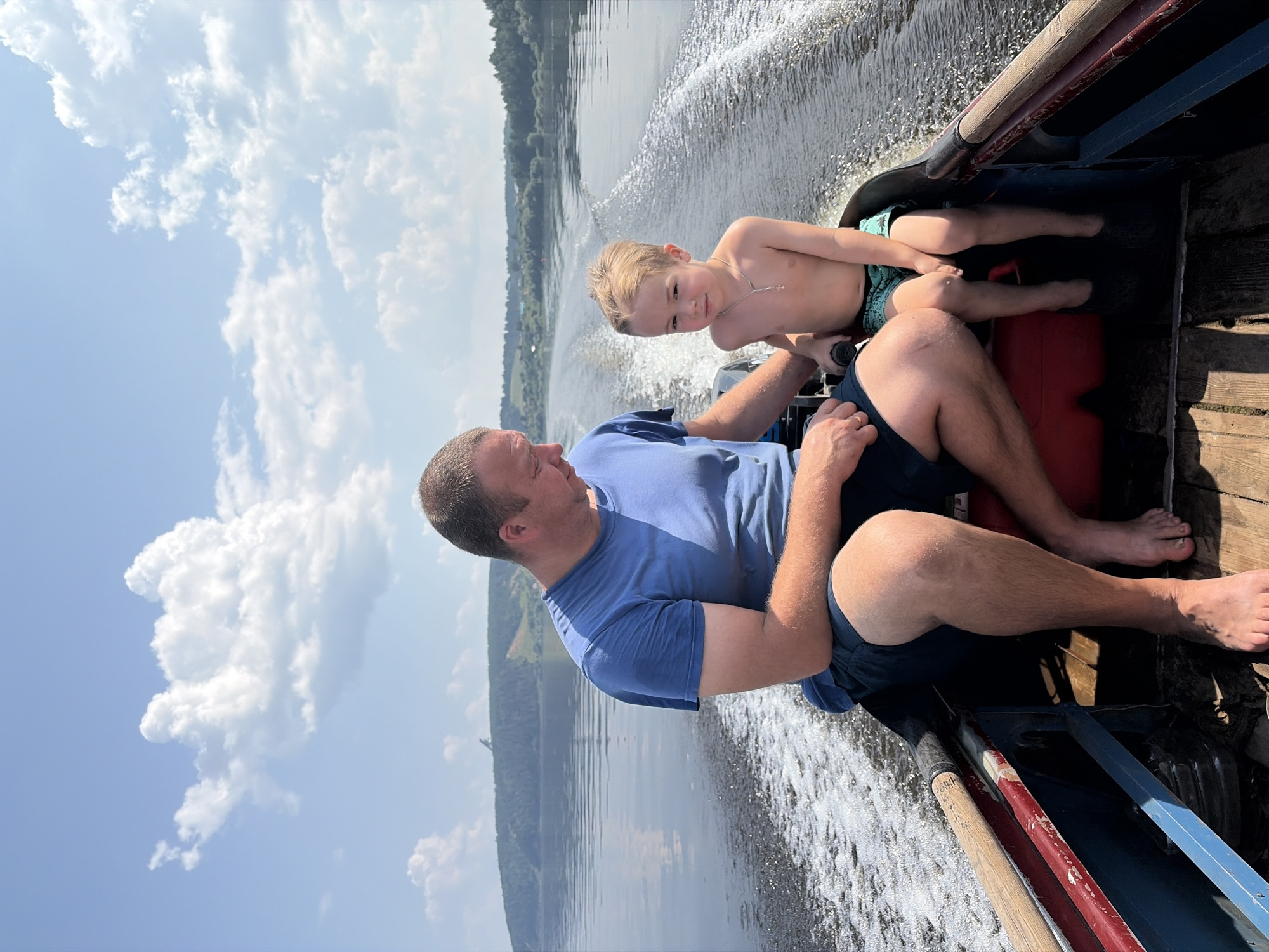 The image depicts a father and son on a boat ride on a river, likely in Ol'khovskoye, Russia, given the context of the request. The father, a man with short brown hair and a blue t-shirt, is seated on the left side of the boat, wearing dark shorts and barefoot. He is looking down, possibly at the boat's controls. Beside him, to the right, sits a young boy with blond hair, wearing only swim trunks. The boy is looking directly at the camera. Both are inside a small motorboat, which is creating a wake as it moves through the water. The river is calm, reflecting the blue sky dotted with white clouds. The background shows a tree-lined shore with a hill in the distance. The boat itself is made of wood, with red and blue accents along its sides, and appears to be a traditional design. The overall scene suggests a peaceful, recreational outing on a sunny day.