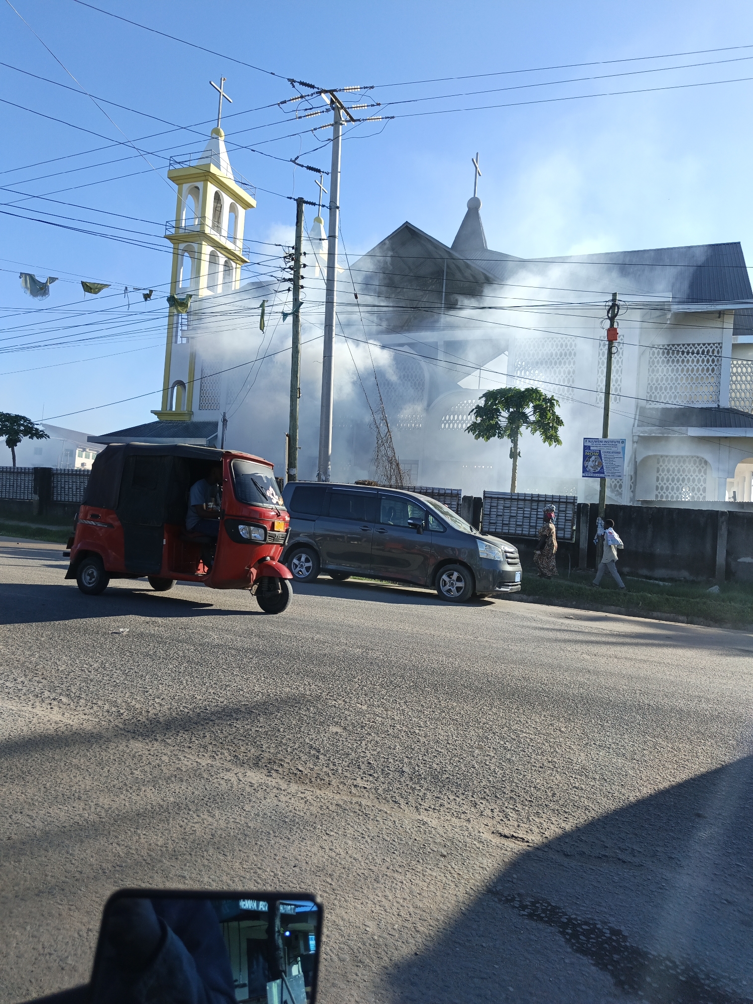 The scene shows a street in Dar es Salaam, Tanzania, with a red auto-rickshaw in the foreground. Behind it, a gray minivan is parked along the sidewalk. Two pedestrians are walking on the sidewalk near the minivan. Smoke obscures part of a large, white building with a cross on top, likely a church, in the background. A yellow tower-like structure with a cross also stands to the left of the church. Utility poles and wires crisscross the sky. The image is taken from a low angle, possibly from inside a vehicle, as evidenced by a rearview mirror visible in the lower portion of the frame.