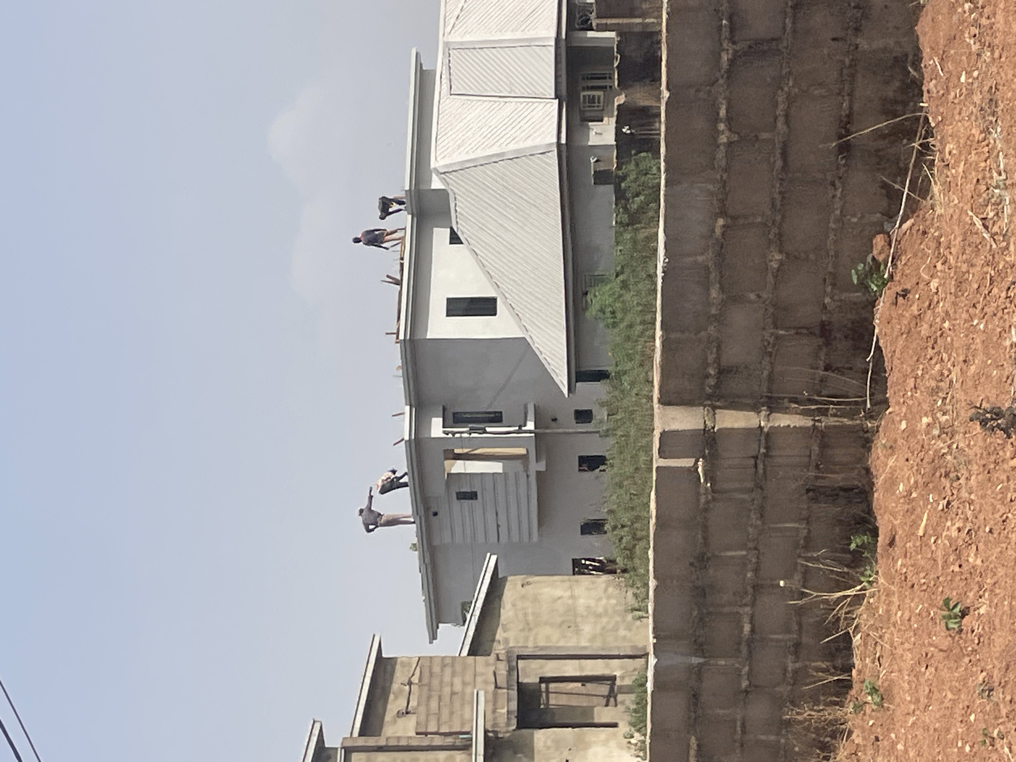 The image shows a building under construction in Jos, Nigeria. Several construction workers are visible on the roof of a two-story building, engaged in what appears to be roofing work. The building has a light-colored exterior, and the roof is made of corrugated metal. A concrete block wall runs along the side of the property, with bare ground and sparse vegetation visible beyond the wall. Another unfinished building is partially visible in the foreground. The sky is light blue and clear. The photo suggests a construction site within a residential or developing area.