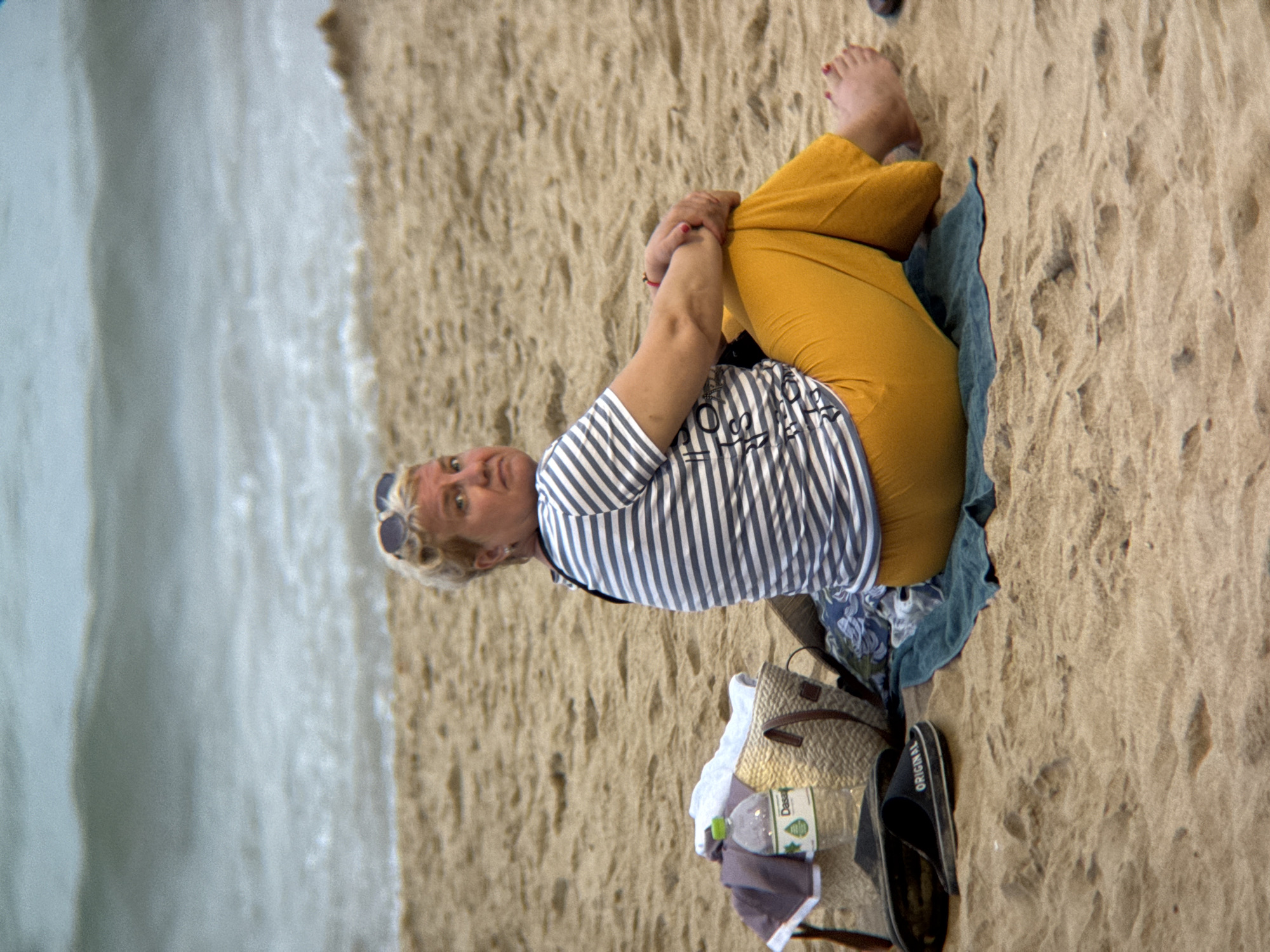 A woman sits on a sandy beach in Nha Trang, Vietnam, facing to her left and looking over her shoulder at the camera. She has blonde, curly hair and is wearing a striped t-shirt and yellow pants. A straw bag, a bottle of water, a pair of flip-flops, and a towel are arranged on the sand next to her. The ocean and waves are visible in the background.