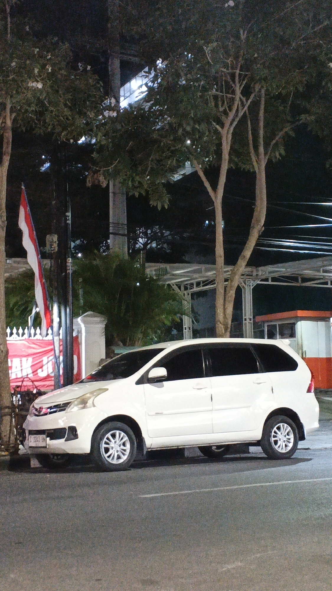 The image shows a white Toyota Avanza parked on the side of a street at night in Bojonegoro, Indonesia. A red and white Indonesian flag is visible on a pole to the left of the car. There is a red banner behind the flag with white text, partially obscured by trees. The car is a multi-purpose vehicle (MPV) with tinted windows and silver alloy wheels. The street appears to be asphalt, with white lane markings visible. In the background, there are trees, streetlights, and some buildings or structures with metal canopies. The overall scene suggests an urban environment at night.