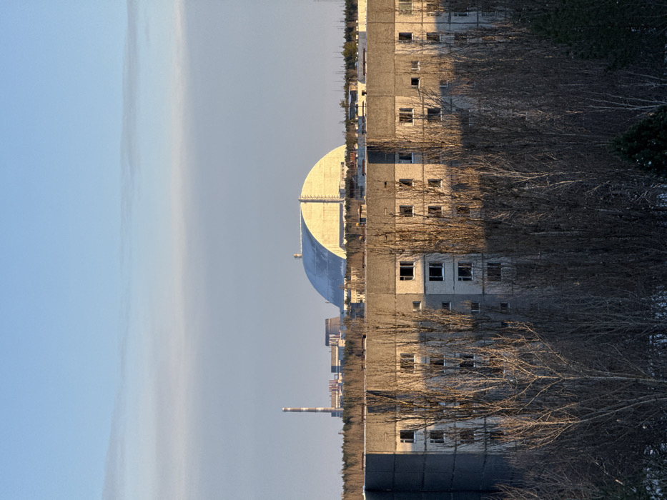 The image presents a panoramic view of Prypiat, Ukraine, looking towards the Chernobyl Nuclear Power Plant complex. The sky is clear, pale blue with faint, high-altitude clouds. In the far background, the large, semi-cylindrical New Safe Confinement (NSC) structure is prominent, partially covered by what appears to be snow or ice on its roof, and illuminated by a low sun from the right. To the left of the NSC, a tall, red and white striped industrial chimney, a dark cooling tower, and other smaller industrial buildings of the power plant complex are visible, situated beyond a dark, dense band of coniferous trees.
In the mid-ground, a multi-story (approximately 5-6 visible floors) concrete panel residential building stands, characterized by its grey facade. Numerous window openings are visible; most appear empty, lacking glass. The upper levels and the right side of this building are illuminated by direct sunlight, casting a warm orange hue, while the lower and left sections are in shadow. The foreground is dominated by a dense arrangement of bare deciduous trees with intricate, leafless branches, largely in shadow, indicative of winter or early spring. Patches of snow are visible on the ground beneath these trees. No individuals, overt actions, or interactions are depicted within the scene. The environment suggests cold weather and a state of abandonment.