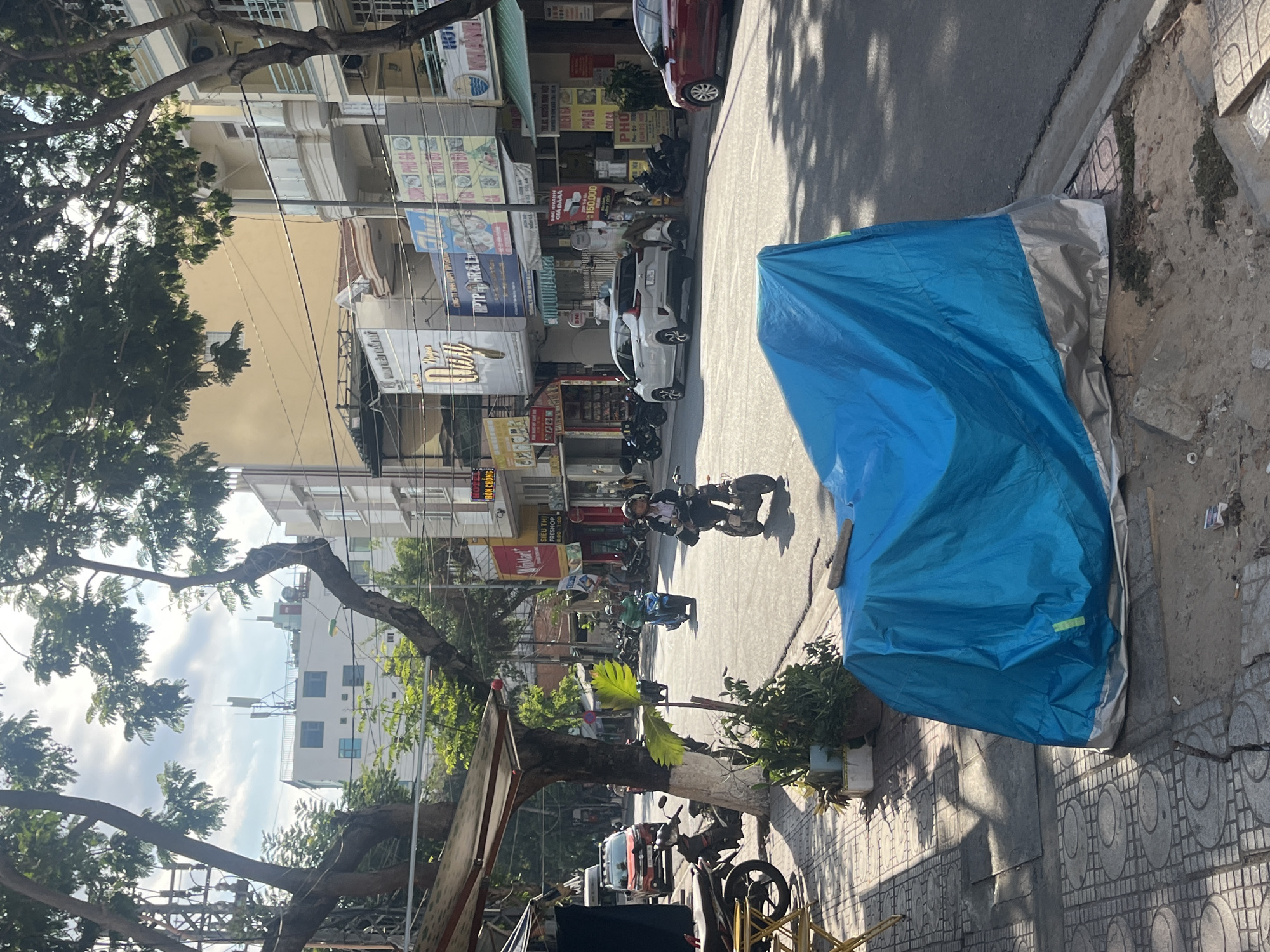 Here's a description of the image:
The image shows a street scene in Bac Nha Trang Ward, Vietnam. A blue tarp covers an object on the sidewalk. The street has vehicles, including cars and motorbikes, and pedestrians. The buildings lining the street have storefronts, including restaurants and shops. A tree stands on the left side of the image, partially obscuring the buildings. Street signage is visible, bearing the names of businesses and prices in Vietnamese currency. The photo appears to be taken in daylight.