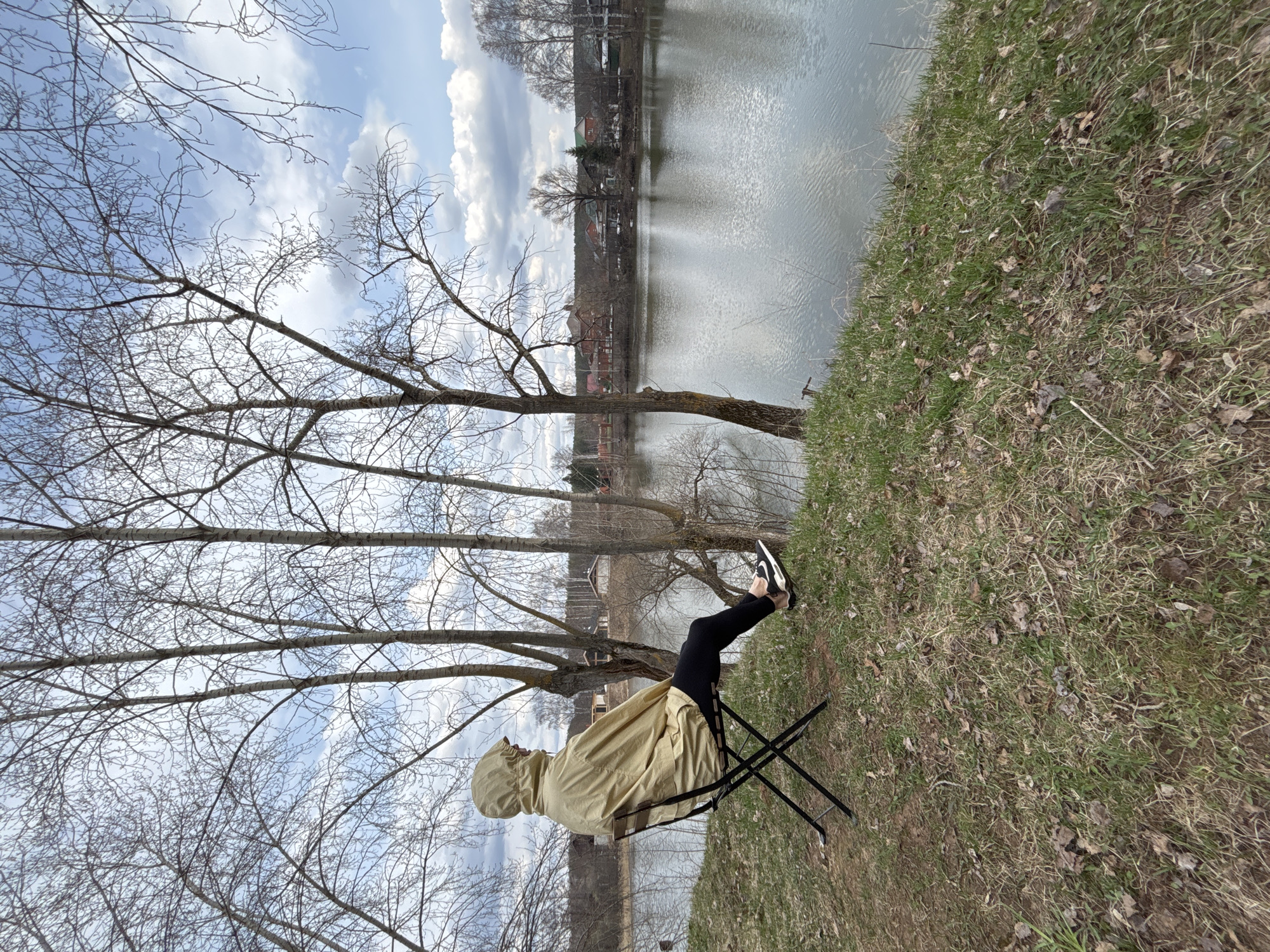 A person sits in a folding chair on a grassy bank next to a body of water, possibly a pond or lake. The person is wearing black leggings, black and white sneakers, and a light brown hooded jacket. The person is facing away from the camera, towards the water. Several bare trees are visible in the foreground, extending up into a partly cloudy sky. The water reflects the sky. Across the water, there are some buildings. The location appears to be Chaikovsky, Russia, based on the presence of buildings and landscape that are common in that city. The photograph is taken at an angle, possibly tilted when snapped.