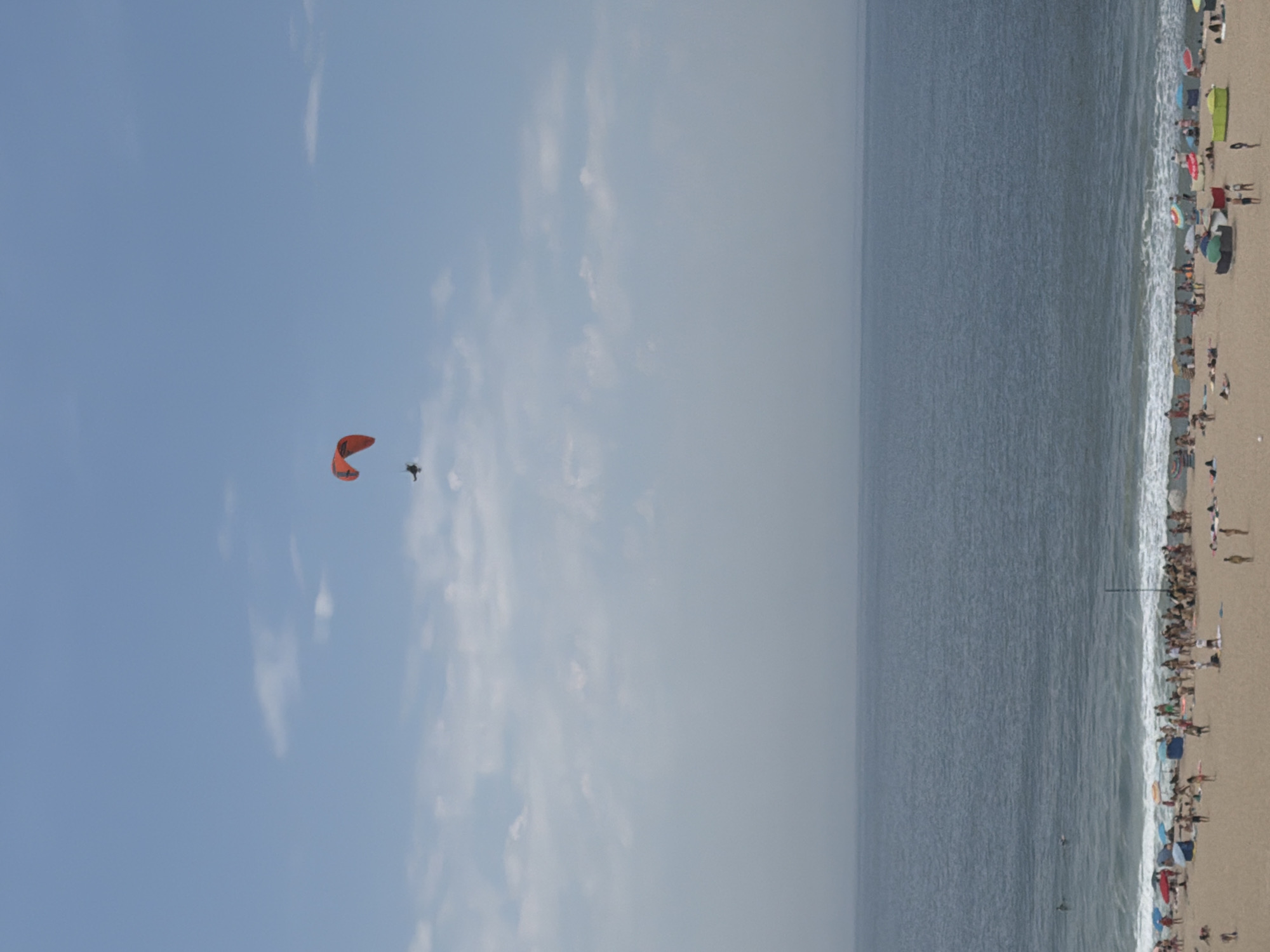 The image shows a sunny day at a crowded beach in Espinho, Portugal. A person is paragliding in the bright blue sky, with the vast ocean to the right. The beach is filled with people sunbathing, swimming, and relaxing under colorful umbrellas. The waves are crashing gently on the shore, creating a peaceful atmosphere. The time of day appears to be late morning or early afternoon, as indicated by the bright sunlight and long shadows. The weather is warm and pleasant, perfect for a day at the beach.