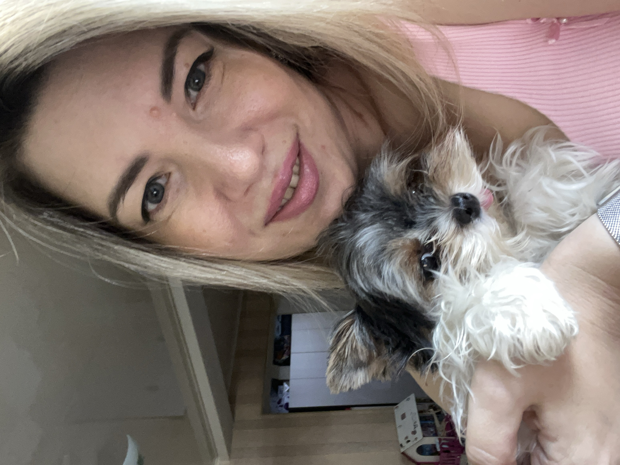 A woman with blonde hair and blue eyes holds a small, fluffy dog with black, white, and grey fur. She is wearing a pink top and smiling. The dog has its tongue sticking out slightly. In the background, there is a light-colored wall and what appears to be a closet with doors. To the left of the woman and dog, there is a small toy house. The image appears to be taken indoors, possibly in a residential setting. There are no identifiable landmarks or indications of the specific location within the city of Sochi, Russia.