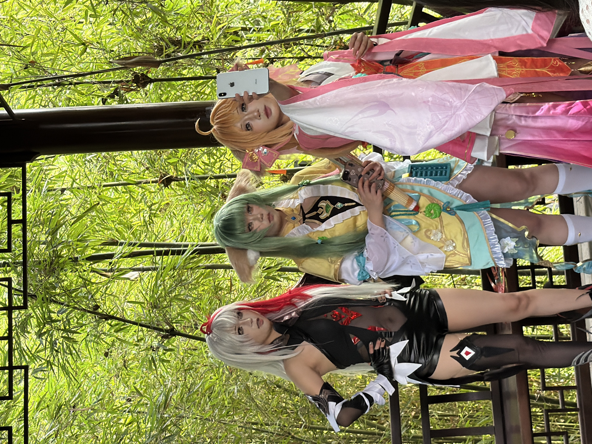 Three individuals in cosplay stand in a bamboo grove. The person on the left has white and red hair and is wearing a black and red costume with black shorts and thigh-high stockings. The person in the middle has green hair and is wearing a yellow and light blue outfit with cat ears. The person on the right has orange hair and is wearing a pink and white outfit. They are holding cell phones, with the person on the right taking a selfie. The background is filled with lush green bamboo stalks. The scene appears to be outdoors, possibly in a park or garden setting within Yuzhong District, China, characterized by traditional architectural elements like the wooden railing and decorative overhead structure.