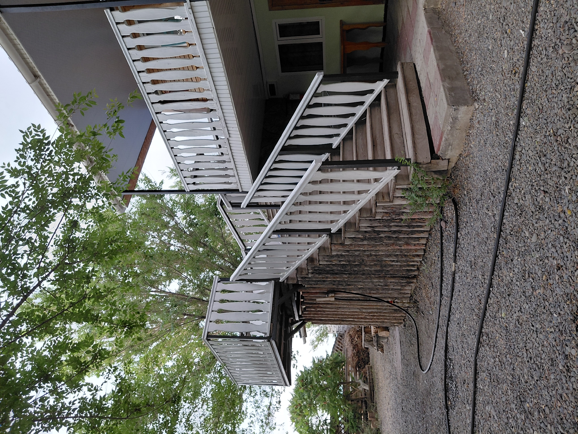This is a tilted, medium-shot photograph of an outdoor staircase leading up to a balcony. The staircase is constructed with wooden steps and sides, with white balusters and handrails. The balusters are a distinctive design, with a wider middle section and narrower ends. A small, covered landing area with similar white balustrades is visible at the bottom of the staircase.
To the left of the staircase, lush green foliage from a large tree is prominent, with branches and leaves reaching into the frame. The tree is dense and leafy, suggesting a temperate climate. The ground surrounding the staircase is covered in small grey gravel. A dark hose is visible snaking across the gravel.
In the background, a building with a light green exterior wall and a dark roof is partially visible. There are at least two windows on this building, one of which appears to be tinted dark. A wooden shelf unit is also visible outside the building. The overall impression is of a rustic, possibly rural, residential setting.
The lighting suggests it is daytime, with diffused light indicating overcast or partly cloudy weather. There are no people visible in the image. The photograph appears to be taken in Ақши, Kazakhstan, as per the provided location context. The tilt of the camera adds a dynamic, slightly disorienting perspective to the scene.