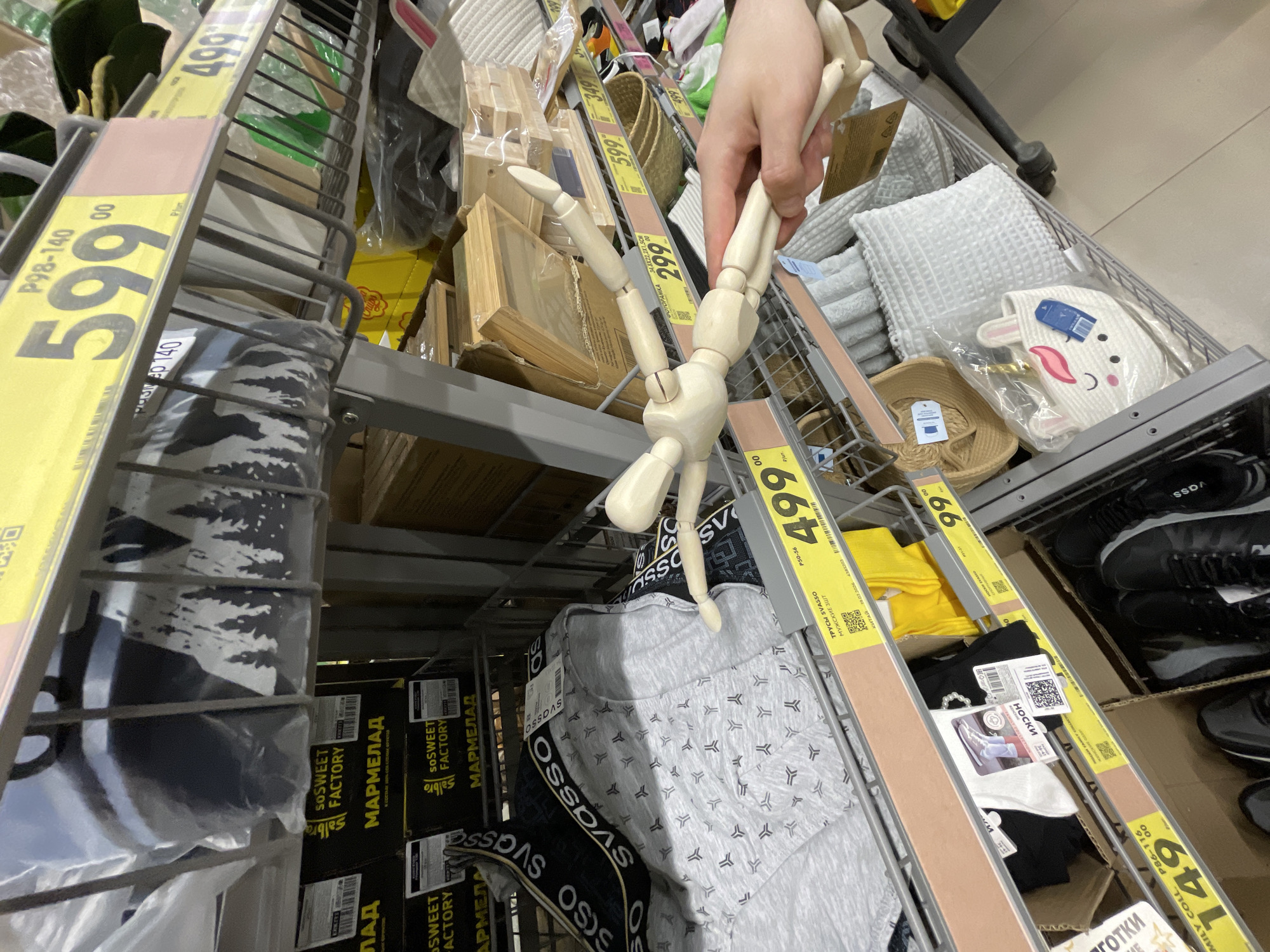 This image shows a shopping cart in Moscow, Russia, containing various items. A person's hand is holding a wooden mannequin doll, suspended above the items in the cart. The cart itself is filled with a variety of goods, including packages of men's underwear ("SVASSO" brand), socks, a decorative rabbit-shaped bag, white waffle weave blankets, small wooden boxes, and woven baskets. Shelving and price tags are visible, indicating prices such as 599 rubles, 499 rubles, and 149 rubles. Products from "SO SWEET FACTORY" ("MARMELAD") are also seen on the shelf.