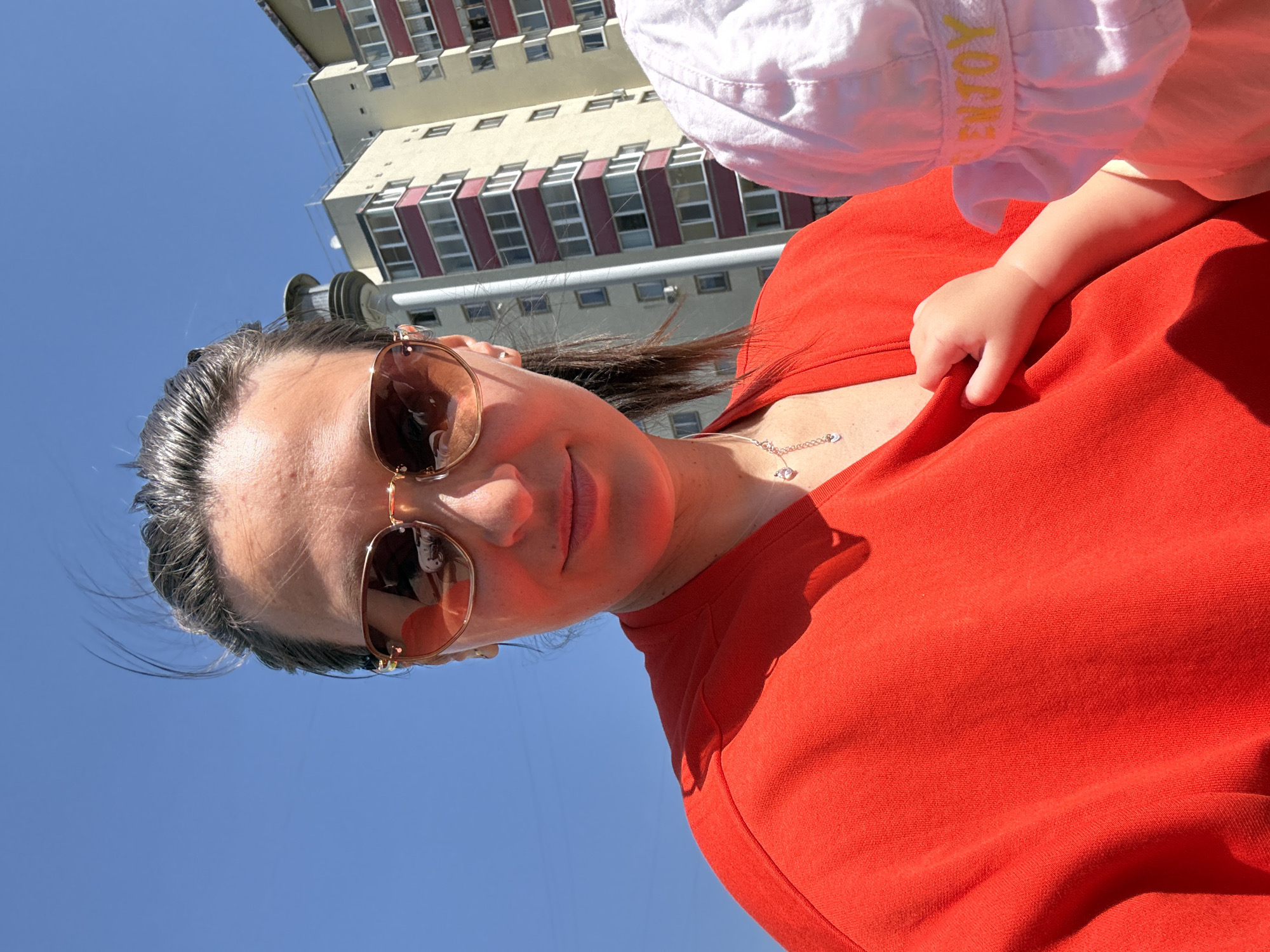 A medium shot shows a woman in the foreground wearing sunglasses and a red shirt. A small child in a white hat is next to her, with their hand grasping the woman's shirt. Behind them is a multi-story building with light-colored walls and maroon window frames. The location is Chaikovsky, Russia, based on the building style. The sky above is clear and blue, indicating a sunny day.