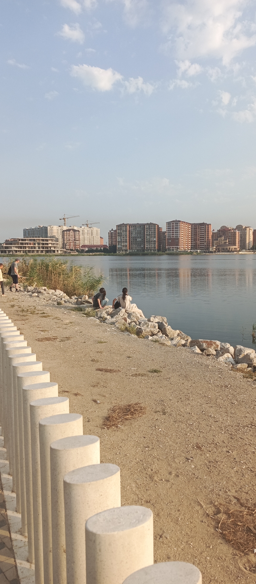 The image captures a waterfront scene in Makhachkala, Russia. In the foreground, a row of cylindrical, light-colored bollards lines a sandy pathway. Beyond the pathway, a rocky embankment separates the shore from a calm body of water.
On the rocky embankment, two individuals are seated, facing the water. One is wearing dark clothing and appears to be looking down, possibly at the rocks. The other is wearing a light-colored garment and is facing away from the camera, also looking towards the water. Further to the left, near some tall reeds, another person in a grey t-shirt and dark shorts is standing, holding what appears to be a fishing rod. Another person in a yellow shirt is partially visible to the far left.
Across the water, a city skyline is visible. Several large, multi-story buildings, some under construction, dominate the background. Two prominent construction cranes are also visible above some of the buildings. The sky is a pale blue with scattered white clouds. The overall impression is a peaceful urban waterfront with people enjoying the view.