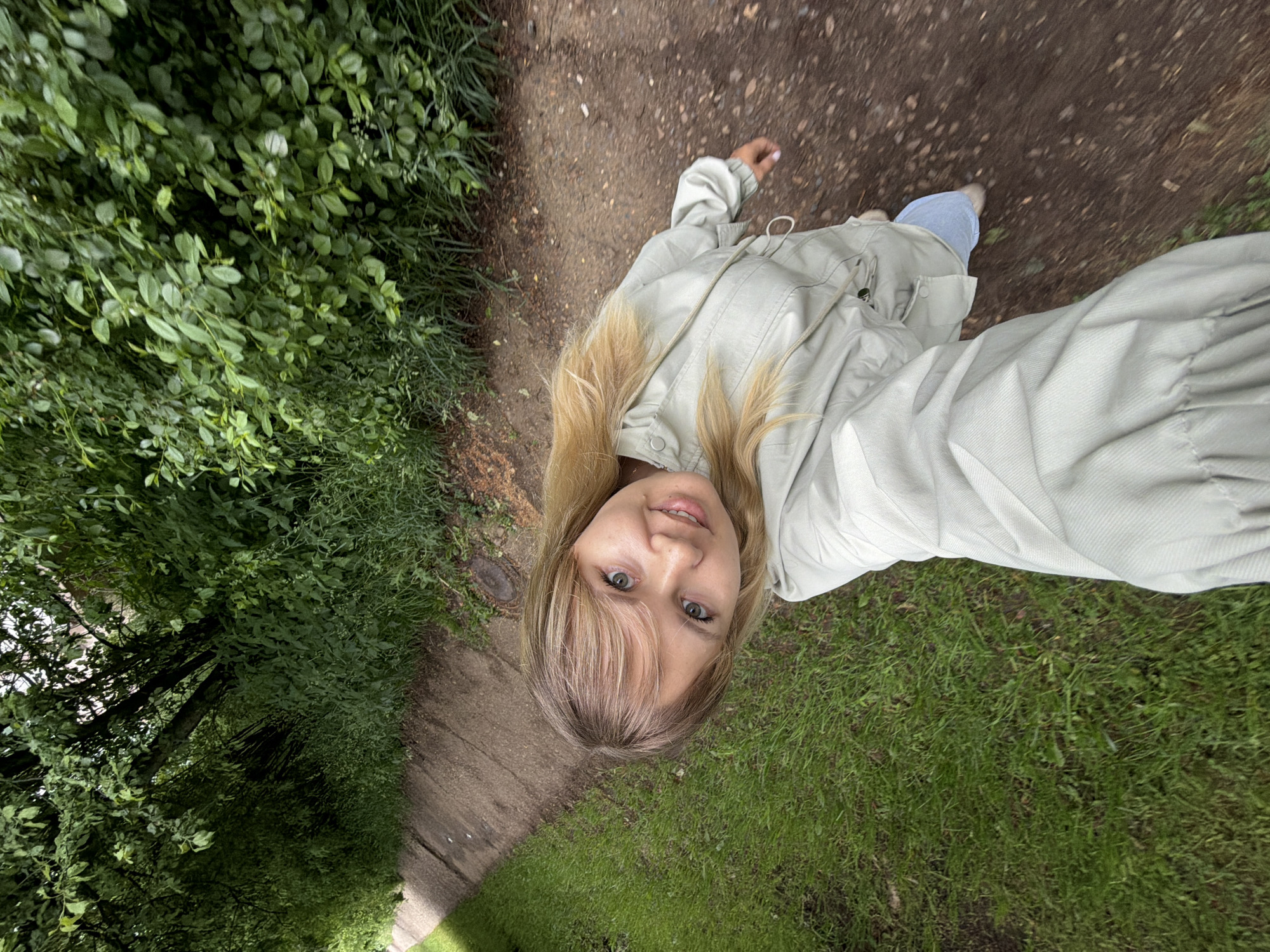 A fair-skinned female with blonde hair is positioned in the center of the frame. She is wearing a light-colored jacket and blue jeans. The background consists of a pathway surrounded by lush green foliage and grassy areas. The image appears to be taken from a high angle, creating a perspective looking down at the subject and the surroundings. The specific location in the city of Chaikovsky, Russia cannot be determined from the provided image alone.