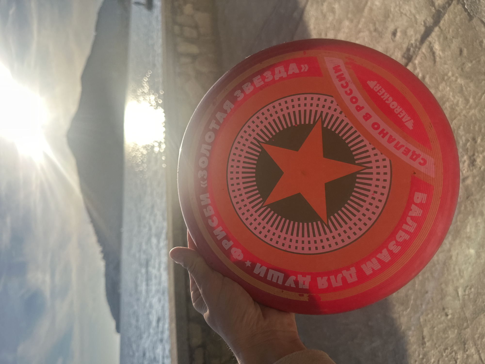 A hand holds a red frisbee with a star and Russian text against a background showing water and a mountain, likely the Adriatic Sea. The frisbee is labeled "Золотая Звезда" (Golden Star). The photo appears to be taken in bright sunlight, creating glare. The location is likely Budva, Montenegro, given the coastal scenery. The frisbee is close up while the other elements are visible further away.