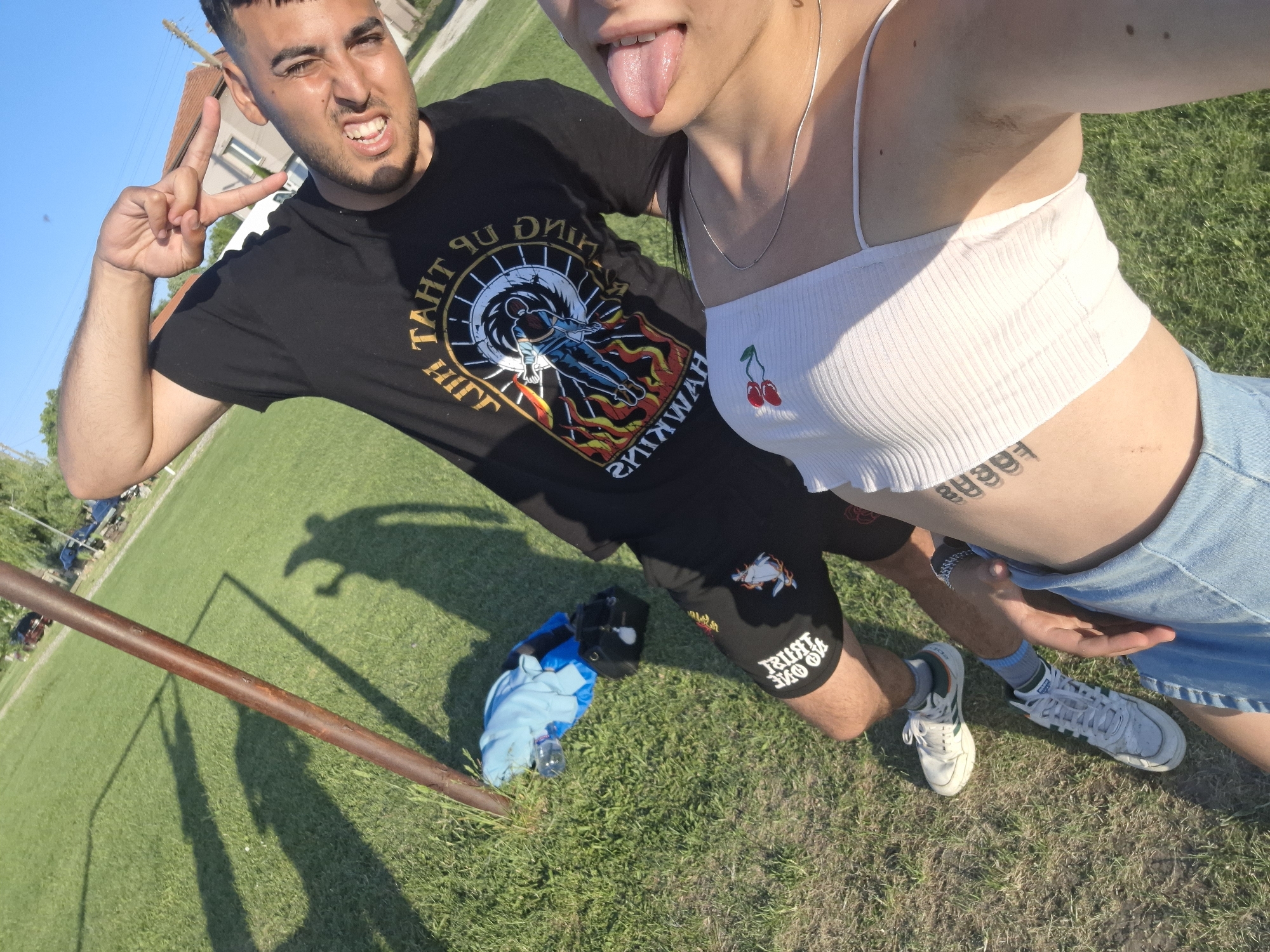 The image shows two individuals on a grassy field in an outdoor setting. A man is on the left, wearing a black T-shirt with a graphic and black shorts with "TRUST NO ONE" printed on the leg. He makes a peace sign with his left hand and displays a facial expression with visible teeth. A woman is on the right, wearing a white ribbed crop top with a cherry detail, a denim skirt, and a silver necklace. She sticks out her tongue. Her abdomen reveals a tattoo. The scene includes shadows cast across the grass, a metal pole is visible on the lower left. Items including a blue shirt, a black cap and a water bottle are also shown on the grass. Based on the image, it is not possible to verify the location in the city of Krapets, Bulgaria.