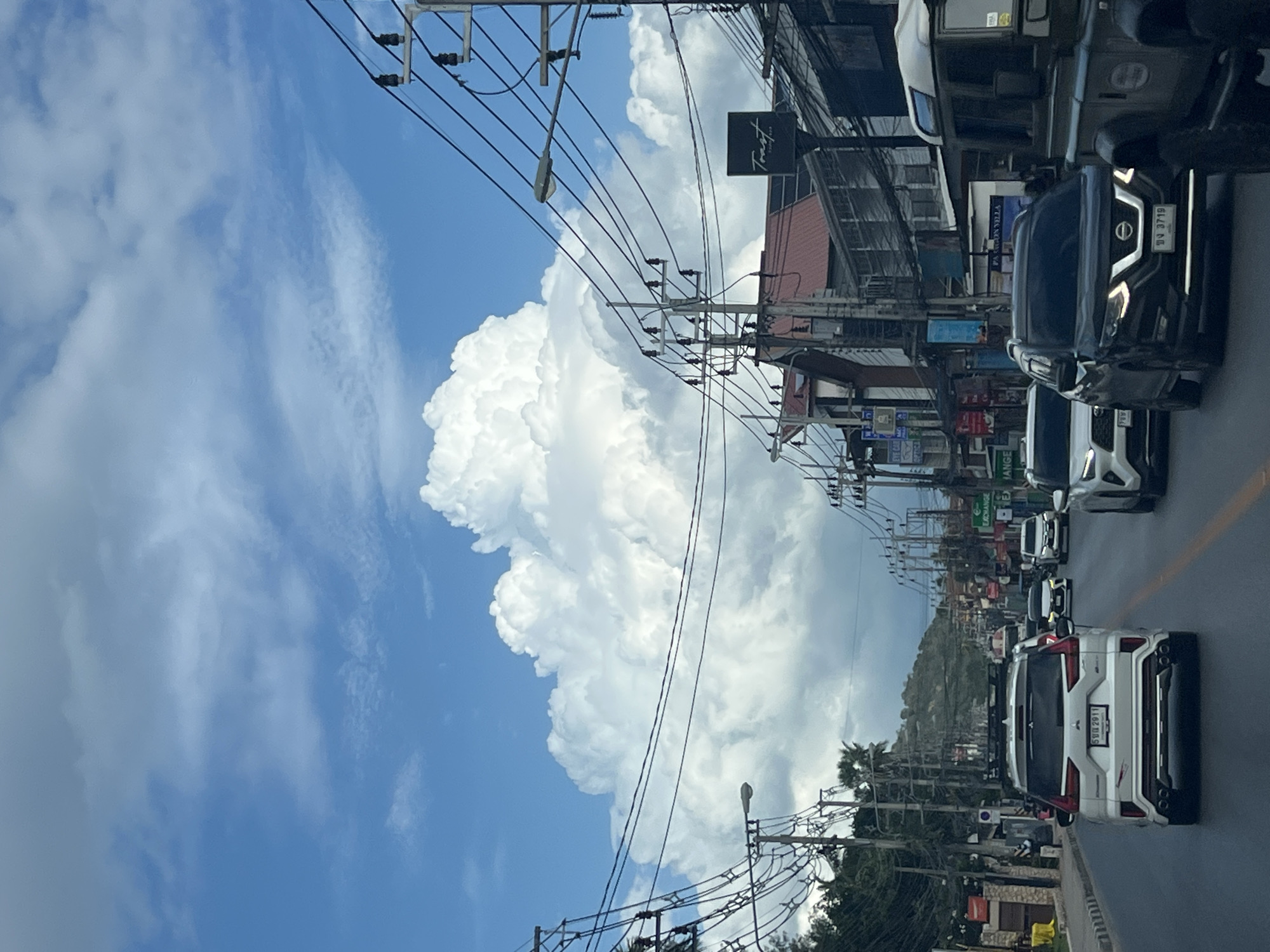 The image shows a view from a car traveling down a street in a city with buildings and utility poles lining both sides of the road. A large, cumulonimbus cloud formation dominates the sky. Several cars are visible on the road, including a white SUV in the foreground with Thai license plates. The buildings appear to be shops or residences, with awnings and signs. The street is paved, and there are sidewalks on either side. The overall impression is of a bright, somewhat overcast day in a tropical or subtropical climate.