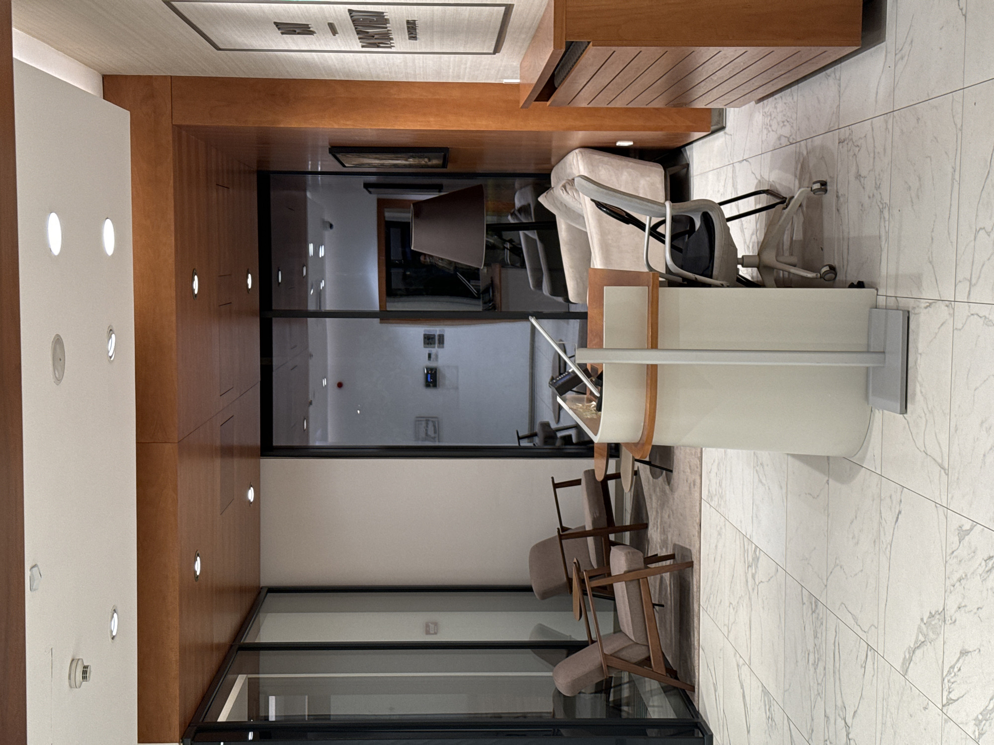 This image shows a reception area of a building, likely an office or business location. The interior design is modern and features wood paneling, glass walls, and a white marble floor. The space includes seating areas with several chairs and a desk or counter area. The lighting is bright. Through the glass walls, additional office areas are visible with desks, chairs, and office equipment.
While the image provides no visual cues directly linking the scene to Belgrade, Serbia, without further information, such as signage or unique architectural elements, it's impossible to confirm the specific location within Belgrade, or even if the location is in Belgrade.