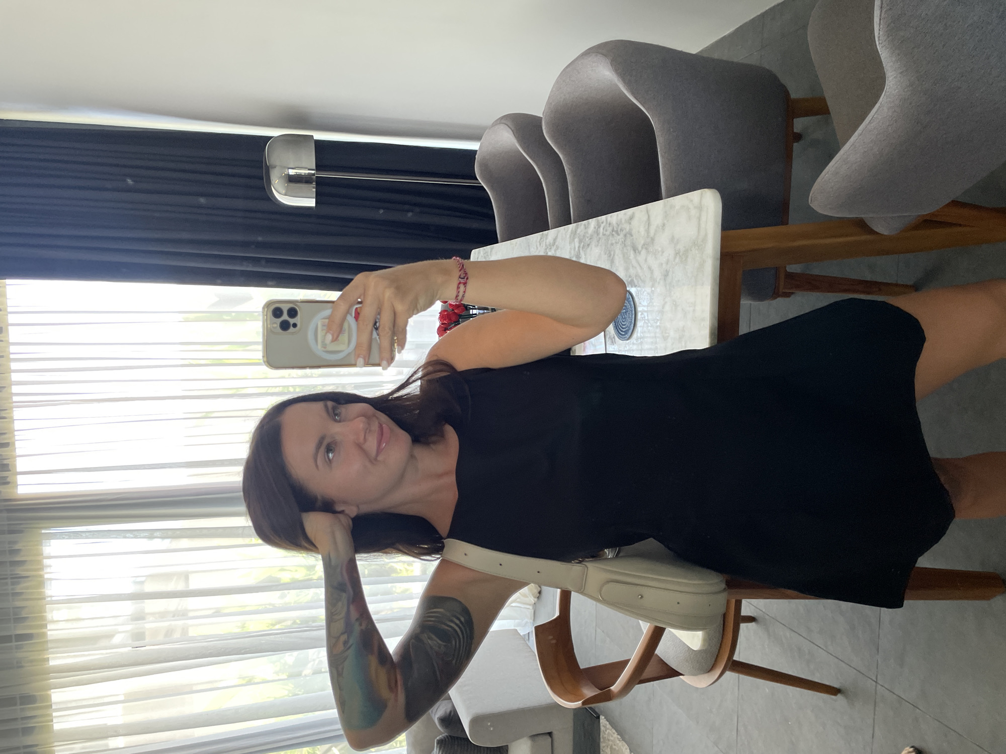 A woman with dark hair and tattoos on her arm is taking a mirror selfie in what appears to be a dining area. She is wearing a black sleeveless dress and a cream-colored shoulder bag. In the background, there is a marble-topped dining table with wooden legs, two grey upholstered chairs, and a floor lamp with a silver shade. A sheer white curtain and a dark curtain are visible on the right. The floor is tiled. The image does not provide any information that would indicate the location is in Kediri, Indonesia.