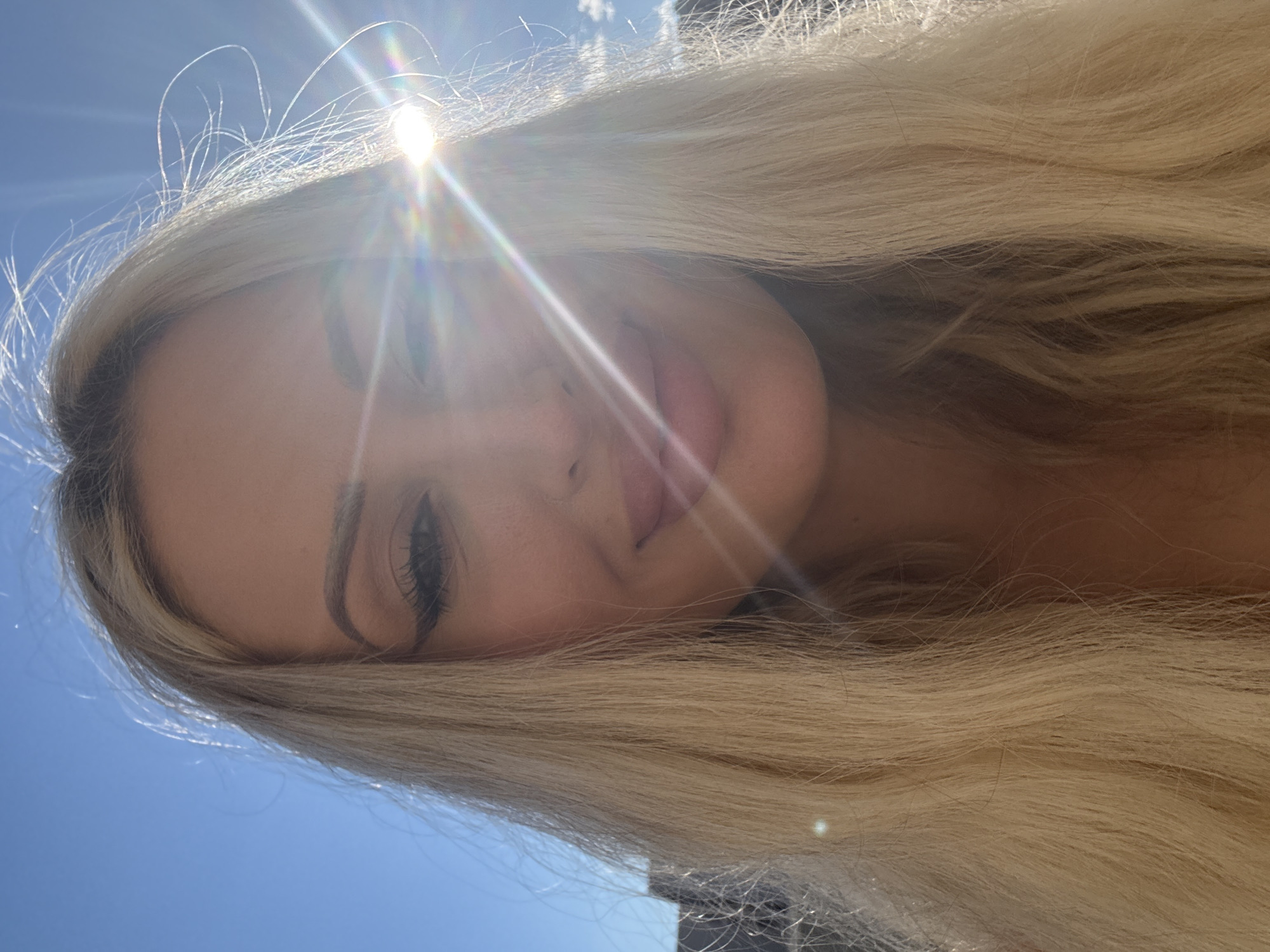 A selfie is taken of a woman's face and upper body, set against a bright blue sky with the sun directly in front of her, creating a lens flare. The woman has long blonde hair that is windswept. She is wearing makeup, including eyeliner and mascara. Her lips are slightly parted, and she is smiling. The background shows a hazy silhouette of a building and some faint clouds. The overall lighting is very bright due to the sun.