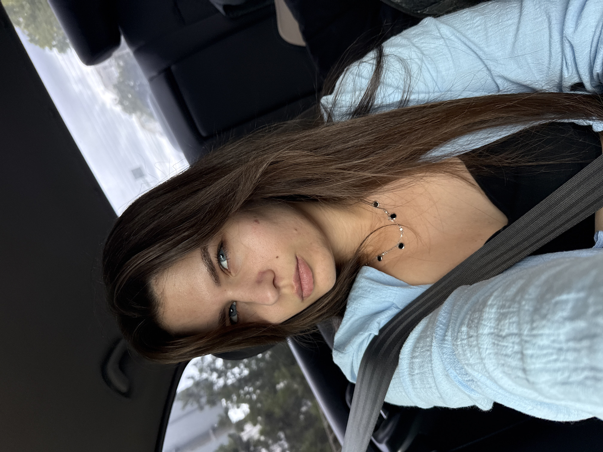 A medium shot shows a woman with long, dark brown hair and blue eyes looking directly at the camera while sitting in the passenger seat of a car. She is wearing a light blue, open-necked shirt over a black top and a silver necklace with black pendants. A gray seatbelt is fastened across her body. The background, visible through the car windows, shows trees and the sky, suggesting an outdoor environment. The interior of the car, including the dark headrest and seat, is also visible.