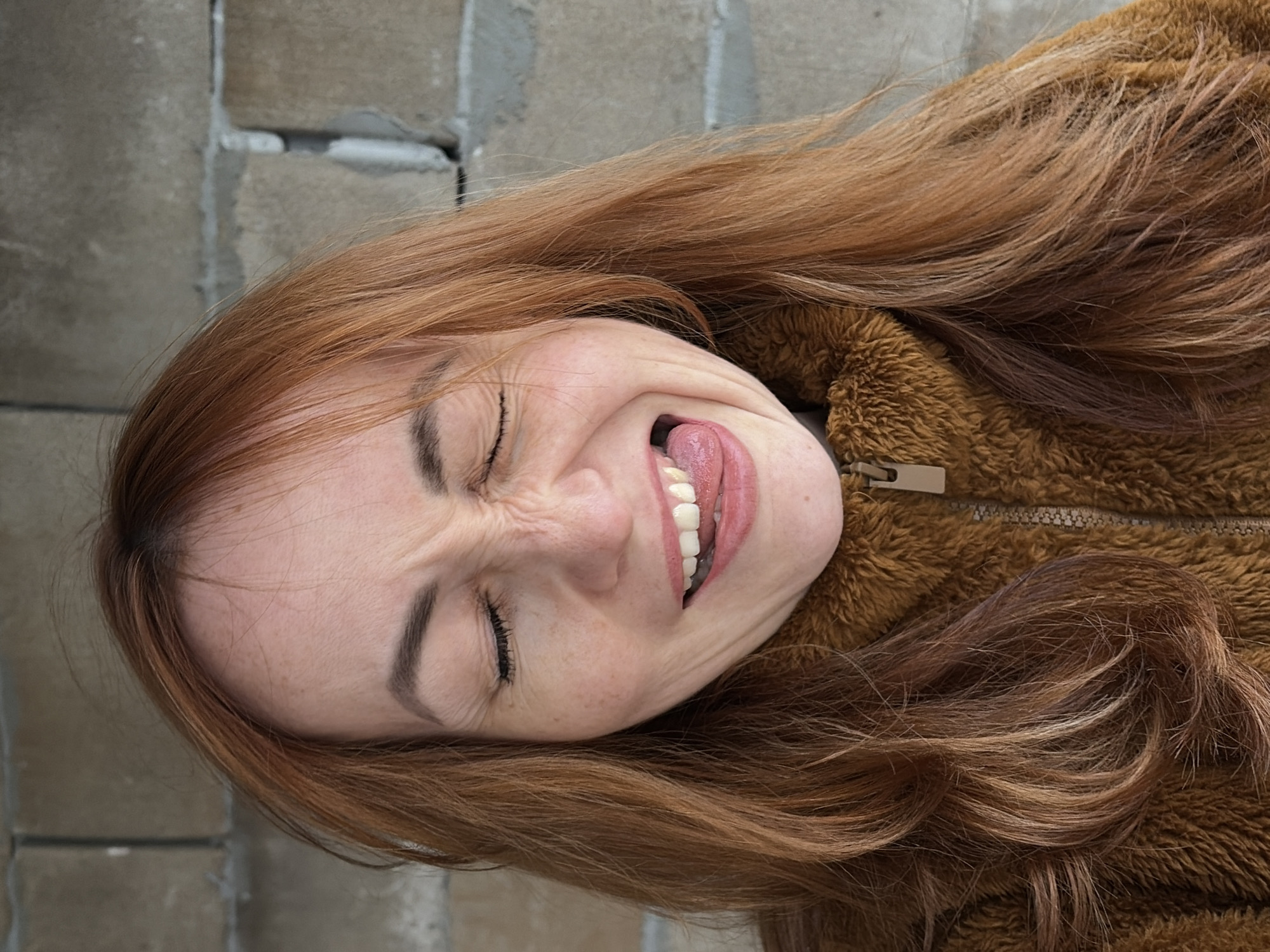 The image features a woman with long, reddish-brown hair, wearing a textured, fuzzy brown jacket with a visible zipper. She is positioned against a background of light grey concrete blocks or bricks, and the photo is rotated 90 degrees clockwise.
The woman is engaged in a playful activity, making a humorous face. Her eyes are tightly closed, causing wrinkles to appear around them, and her face is scrunched up. She has her mouth open, with her tongue extended slightly and visible teeth. This expression conveys a sense of silliness, joy, and lightheartedness. Her hair cascades around her face and over her shoulders, resting on the brown jacket. The lighting appears natural and bright, suggesting it is daytime. There is no visible text in the image. The setting is in Chaikovsky, Russia, though the background offers no specific local landmarks.