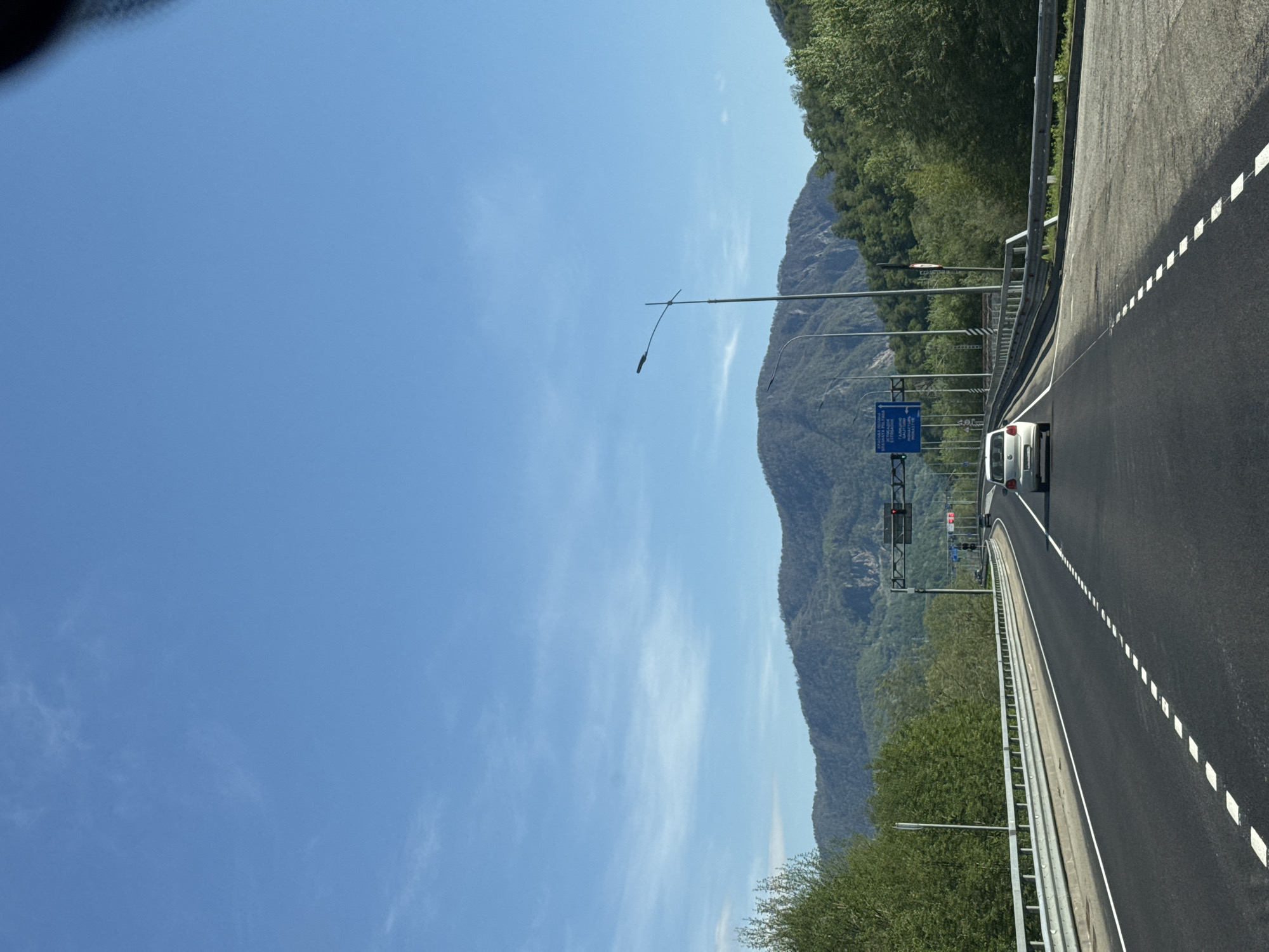The image depicts a road in Monastyr', Russia. A white car travels on the right lane of the road. A mountain covered with trees runs parallel to the road on the left. Street lights line the left side of the road and the sky is clear and blue.