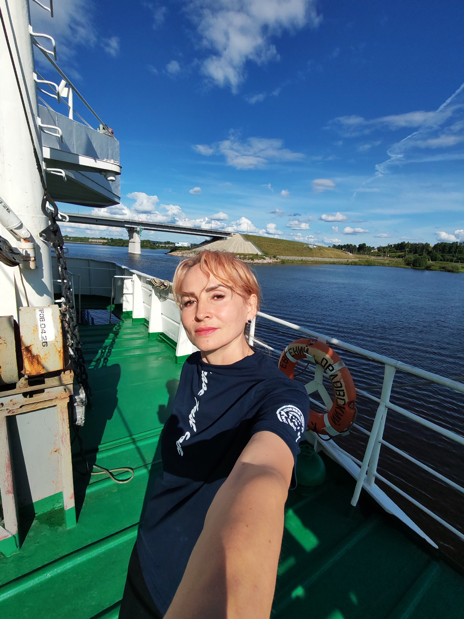 An outdoor, eye-level, medium selfie shows a fair-skinned woman with short, blonde hair and pink lipstick posing on the deck of a boat. She is wearing a dark blue t-shirt with white text and a circular emblem on the left sleeve. Her right arm is extended, holding the camera. Behind her, to the left, is a white railing with some metal equipment and chains. The green deck of the boat is visible in the foreground. To the right, a life preserver is attached to the white railing. The background features a wide, blue body of water, a bright blue sky with scattered white clouds, and a distant bridge with a concrete embankment and trees. The environment suggests a sunny day in the city of Sheksna, Russia, possibly on the Sheksna River.