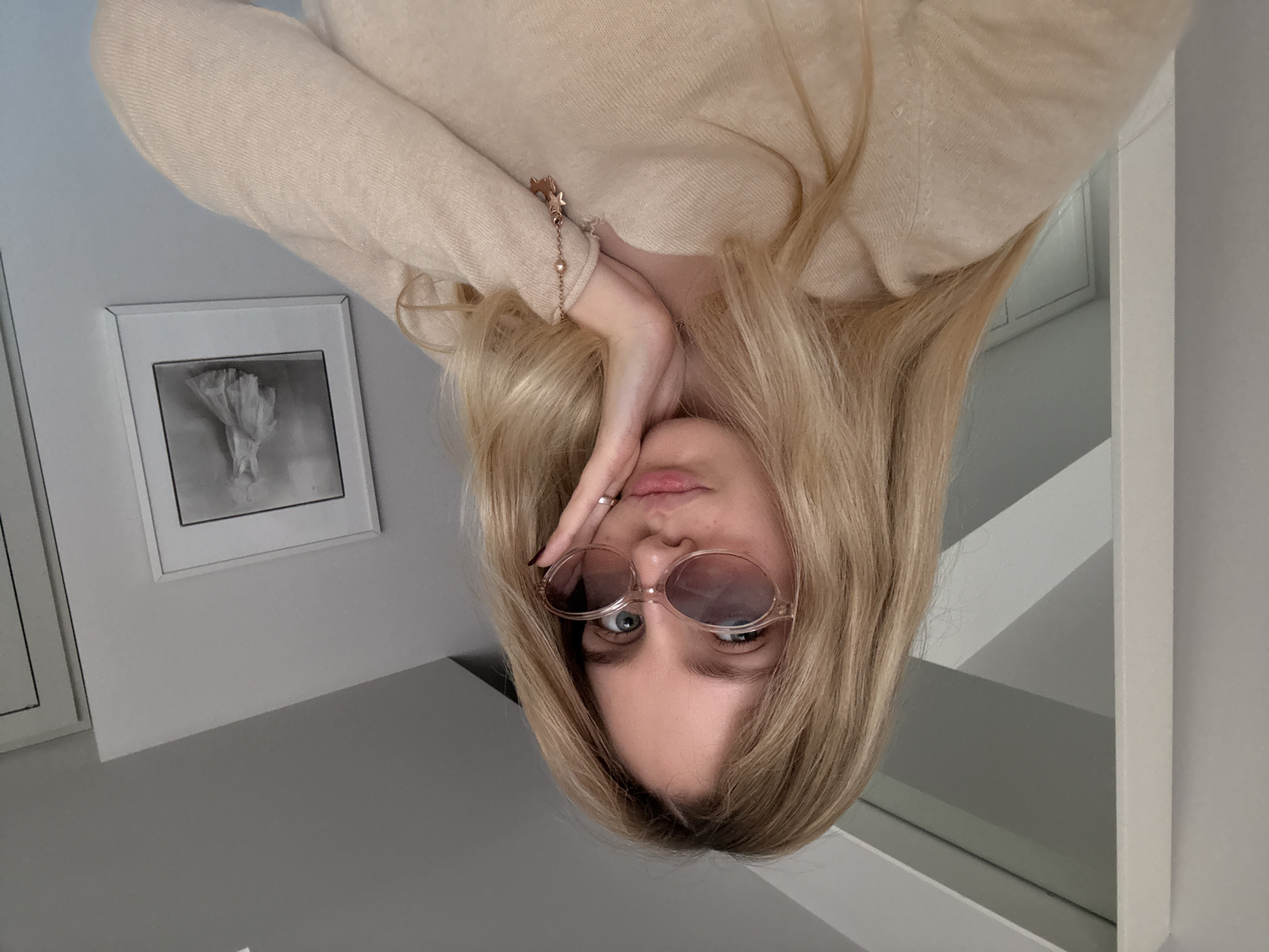 A blonde woman wearing a beige sweater and sunglasses poses for a selfie. She is indoors, standing against a gray wall with framed artwork. The photo is taken upside down.