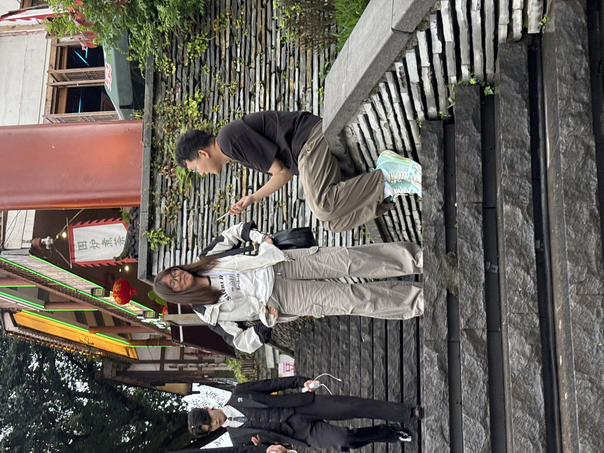 The image depicts a group of people on a street in what appears to be Shapingba District, China. A man in a black t-shirt and brown pants is leaning against a stone wall, looking at his phone. A woman wearing a white jacket and tan pants stands next to him, smiling at the camera. Another person dressed in a black suit and vest is walking in the opposite direction, holding a white device. In the background, there is a traditional Chinese building with red lanterns and green neon lights. The surroundings suggest an urban environment with stone structures and greenery.