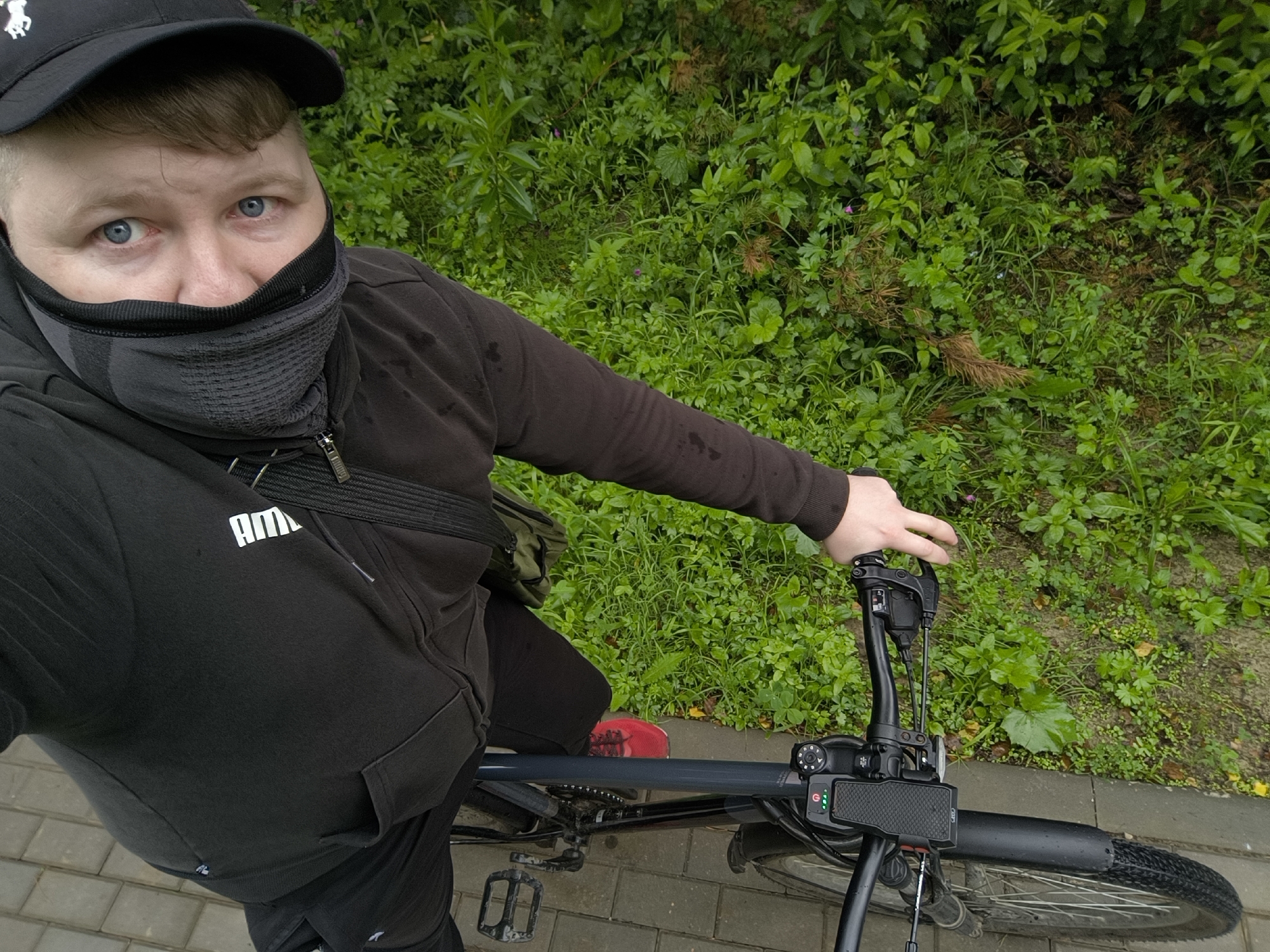 In an overhead shot, a person wearing a black baseball cap, a dark gray neck gaiter pulled up over their nose and mouth, and a black Puma hoodie is seated on a bicycle, holding the handlebars. The person's blue eyes are visible above the neck gaiter. The bicycle has a black frame and handlebars, with a display unit mounted on the handlebars. A black bag with a strap is worn across the person's chest. The background consists of lush green foliage and a paved walkway. The image is tagged with the location of Tarusa, Russia, suggesting the scene takes place within the city limits.