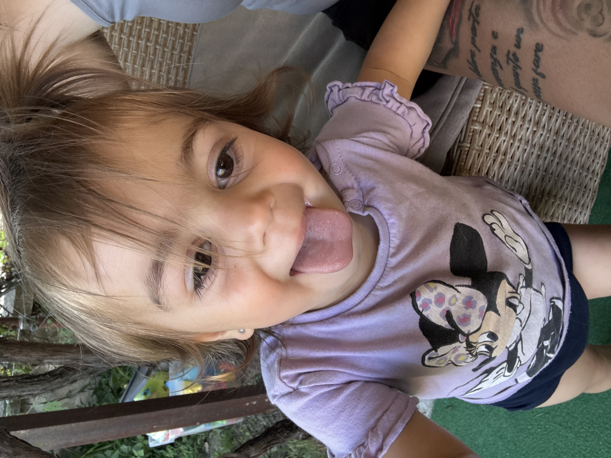 A young child with light brown hair and brown eyes sticks their tongue out. The child is wearing a light purple t-shirt with a Minnie Mouse graphic and dark blue shorts. The child is seated outdoors, with a wicker chair visible behind them and a wooden fence and greenery in the background. The image appears to be taken in Oreshak, Bulgaria, indicated by a tattoo on an adult's arm which is partially visible, bearing text in Bulgarian. The tattoo reads "Shto nosi..." which is an excerpt from a poem by Hristo Botev, a prominent Bulgarian revolutionary and poet, whose birthplace is Oreshak.