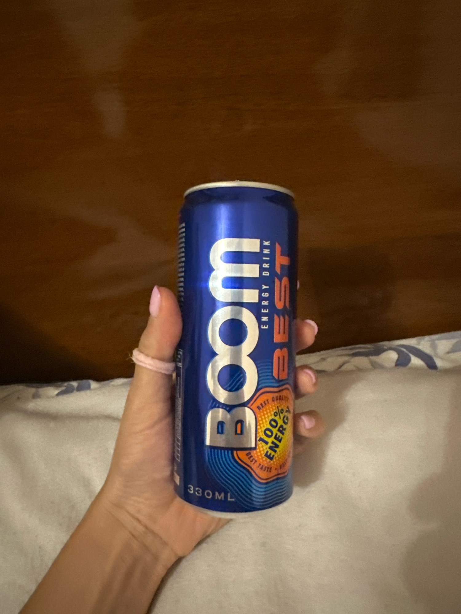 A hand, adorned with light pink nail polish and a light pink hair tie around the thumb, holds a blue can of "BOOM ENERGY DRINK BEST". The can features prominent white lettering for "BOOM" and orange lettering for "BEST ENERGY DRINK". A circular emblem on the can reads "BEST QUALITY 100% ENERGY BEST TASTE". The hand is positioned in front of a white, patterned fabric surface and a wooden headboard. The overall scene is likely indoors. There is no identifiable location in the city of Gyumri, Armenia present in the image.
