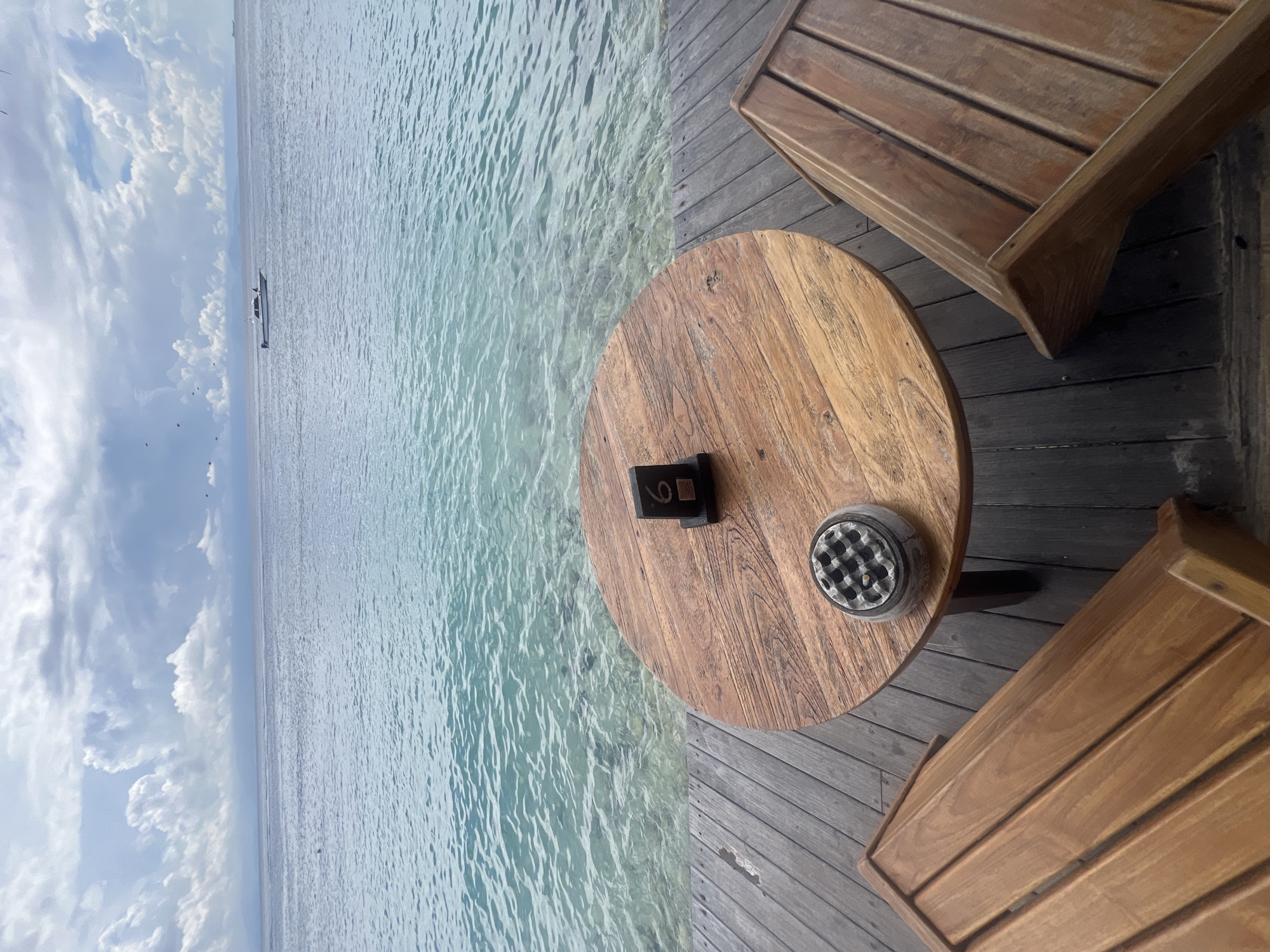 This image captures an outdoor dining or lounge area set on a wooden deck overlooking a calm body of water, likely in Nusa Penida, Indonesia.
In the foreground, a round wooden table takes center stage on the right side of the frame. On the table, there is a black stand displaying the table number "6" and a round, silver-colored ashtray. Partially visible wooden chairs with slatted backs are positioned on the wooden deck around the table, suggesting a seating arrangement for two or more people. The deck itself is made of dark wooden planks.
The left side of the image is dominated by clear, turquoise water, which exhibits gentle ripples and reveals the sandy or rocky seafloor beneath, indicating shallow depths. The water extends towards the horizon, where a small boat can be seen in the distance. Above the horizon, the sky is bright with scattered white, fluffy clouds.
The scene depicts a peaceful and relaxed atmosphere during what appears to be daytime, likely late morning or early afternoon, given the natural light and the clarity of the water. The setting suggests a tranquil coastal or island location perfect for leisure.