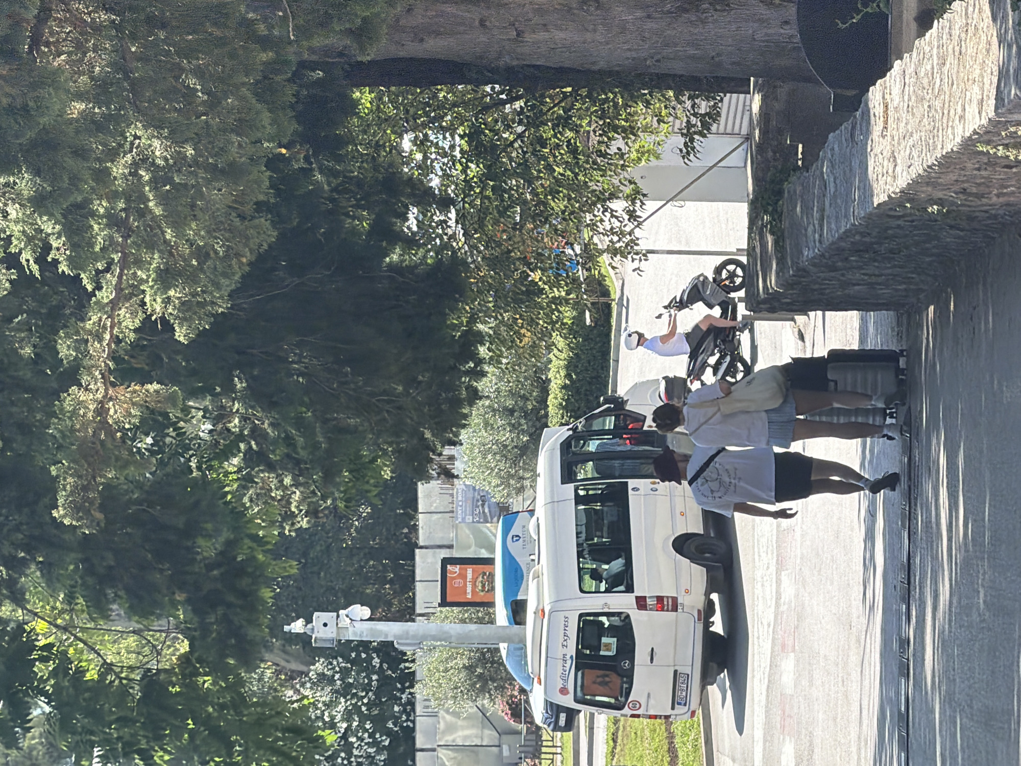 A street scene in Budva, Montenegro, shows people and vehicles on a sunny day. In the foreground, a white minivan with "Mediterranean Express" printed on its rear is parked. Beside it, a person wearing a white t-shirt and black shorts walks away from the camera, with a dark bag slung over their shoulder. Further down the street, a group of people stand near the minivan.
In the background, another white minivan and a blue bus are visible. A person on a scooter rides past, and other individuals are seen in the distance. The scene is bordered by a stone wall on the right and lush green trees on the left. The sunlight casts distinct shadows on the ground, indicating it is likely midday. The overall mood appears casual and relaxed, suggesting a typical day in a tourist area. There are signs in the distance, but they are not clearly legible. The license plate on the white minivan reads "BG 8TB45".