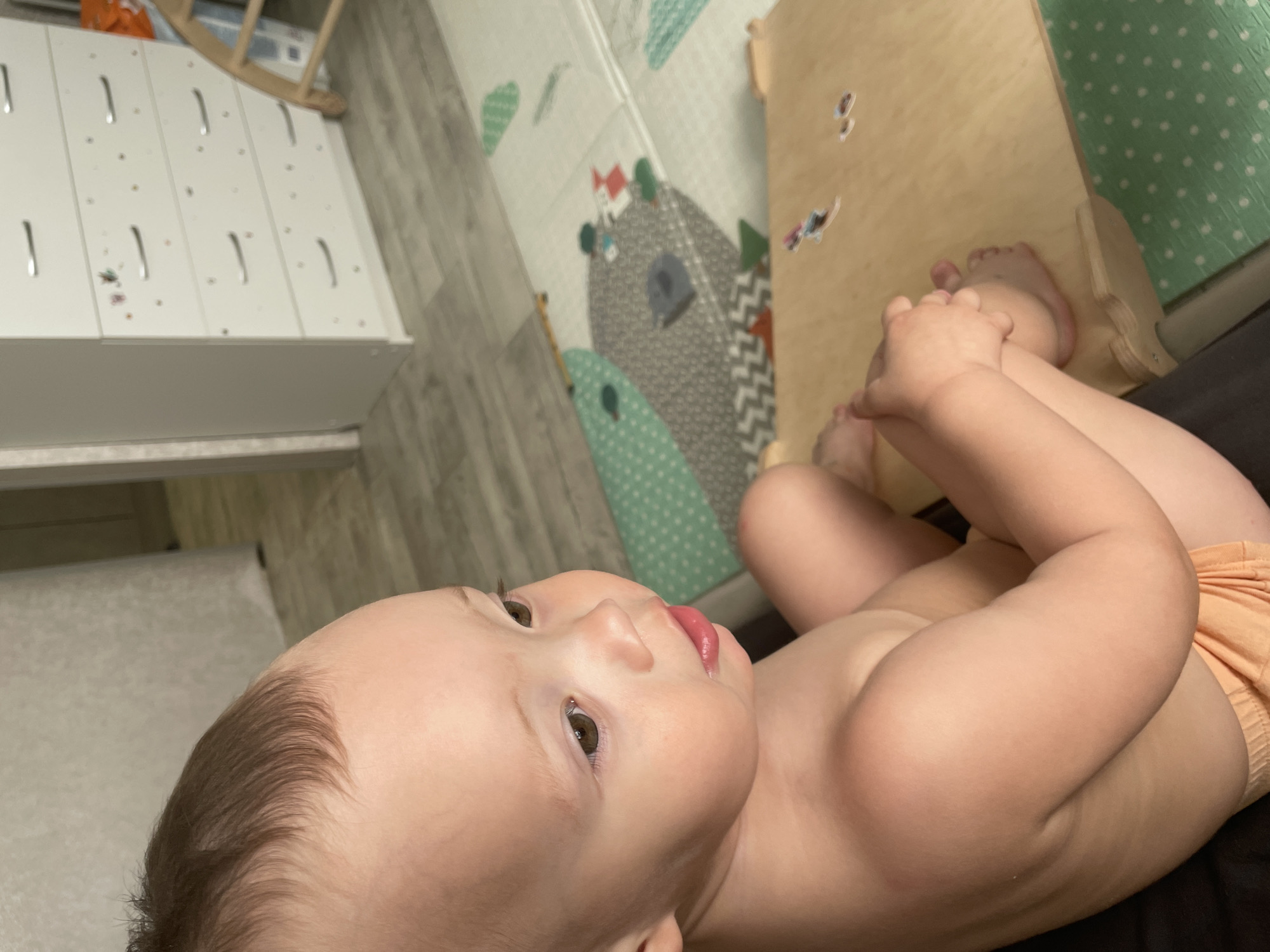 A baby lies on their back. Their head faces to the left. The baby’s legs are raised and they are holding their feet. The baby wears a diaper. A play mat with animal designs is on the floor. A white cabinet is visible in the background. It is not possible to determine the location as Perm, Russia from the image.