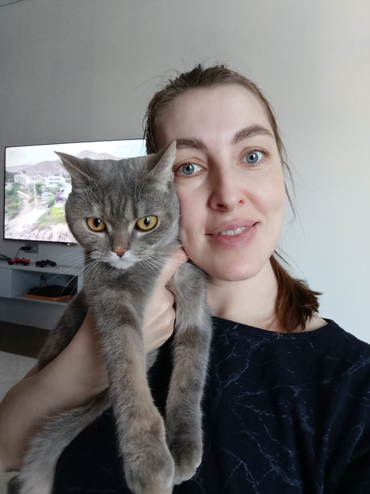A woman and a gray cat are in the frame. The woman is fair-skinned with blue eyes and light brown hair pulled back in a low ponytail. She is wearing a dark, possibly marble-patterned, top. She is smiling slightly and holding the cat. The cat is gray with yellow eyes and appears to be a British Shorthair or similar breed. In the background, there is a flat-screen television mounted on a wall. Below the television, there is a white console shelf with what appears to be game consoles and controllers. The image on the television screen depicts a landscape with buildings, possibly a residential area. Based on the architectural style and landscaping, the location could be in a coastal or hilly region. The room is lit with natural light, giving a bright and airy feel. No evidence suggests that the location is within the city of Chaikovsky, Russia. The information about the location within the city of Chaikovsky, Russia is incorrect.