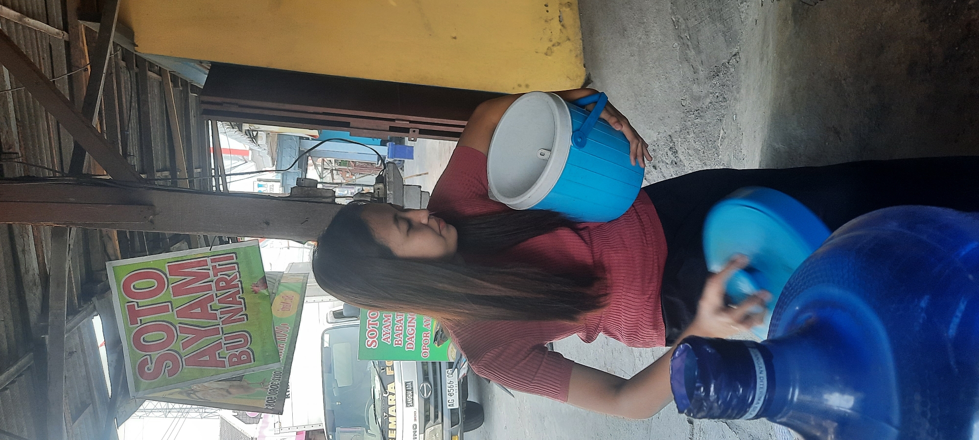 In Selopuro, Indonesia, a woman with long, dark hair and a red, ribbed t-shirt is holding a blue insulated container and a blue lid. She is standing in front of a yellow wall and a truck with a sign that reads "SOTO AYAM BU NARTI". There are two large blue water dispensers in the foreground, one of which the woman is interacting with the lid. The environment appears to be outdoors, possibly a street or market. The lighting suggests it is daytime.