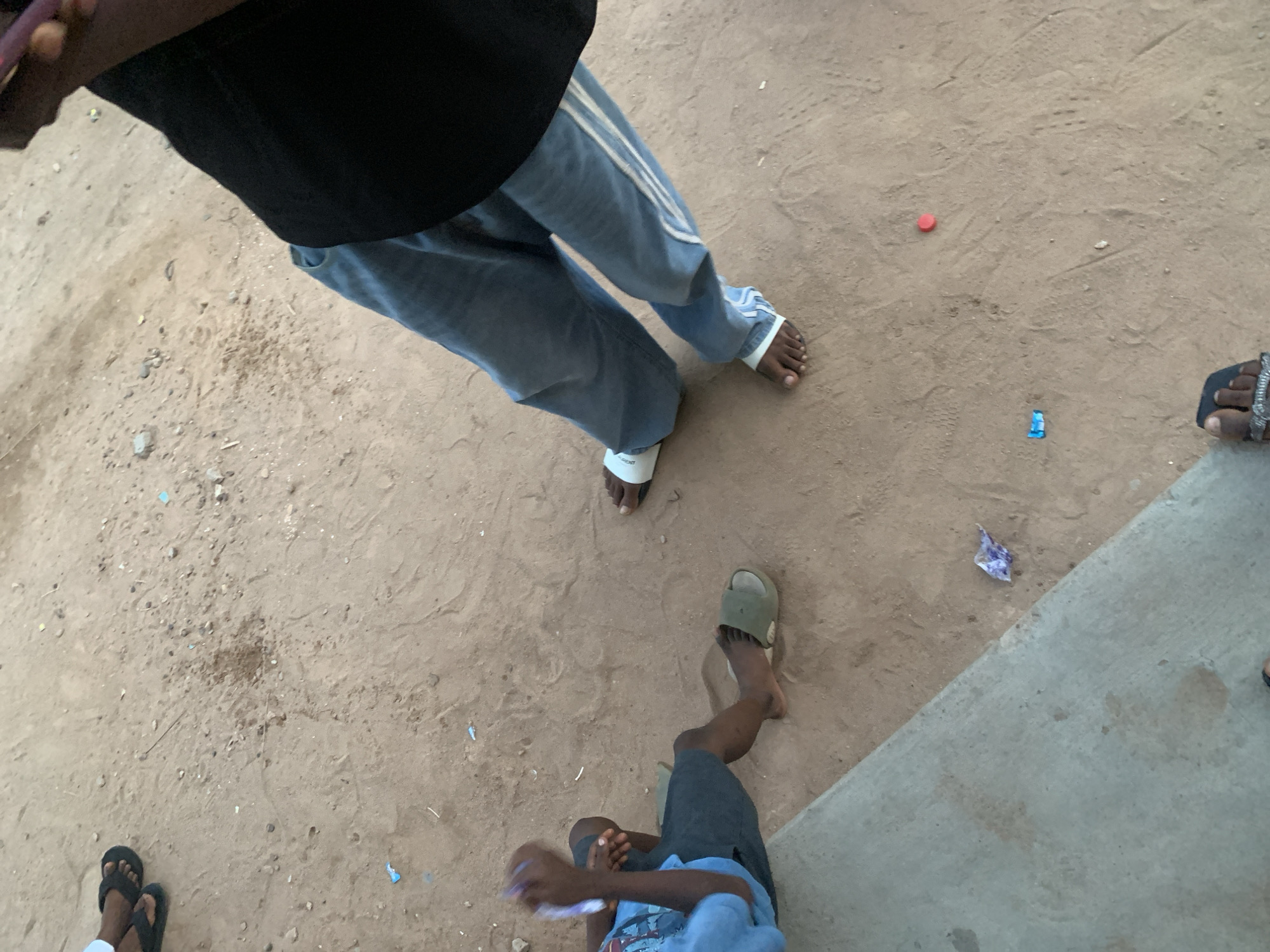 The scene shows an outdoor dirt area in Lokoja, Nigeria. Four people are partially visible from an overhead perspective. One person wears a black shirt, light blue jeans with white stripes, and white sandals. Another wears an olive green sandal and grey shorts. Another has on a black sandal. There are scattered items on the ground, including a red bottle cap and a small piece of trash. A corner of a concrete structure is visible on the right side of the frame.