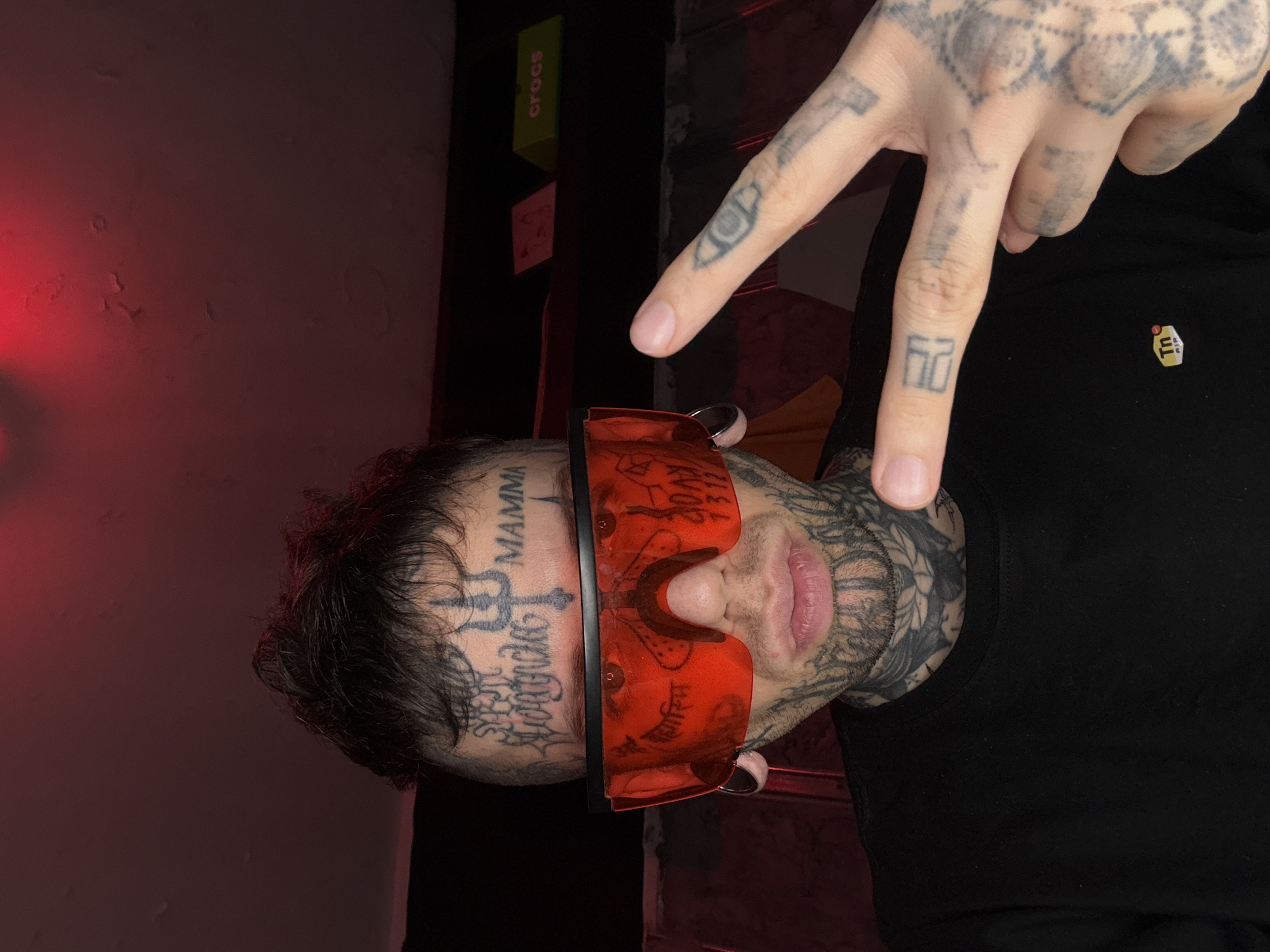 A male individual, heavily tattooed, is captured in a chest-up portrait shot, looking directly at the camera. He has dark, short hair and wears oversized, red, wrap-around sunglasses. Numerous tattoos cover his face, including "MAMMA" on the right forehead, a trident symbol centrally on the forehead, and various scripts and symbols across his forehead, cheeks, and neck. Large, silver-colored circular ear gauges are visible in both ears. He wears a plain black long-sleeved t-shirt, featuring a "Tn AIR" logo on the left chest. His right hand is raised, forming a V-sign with the index and middle fingers, which are also extensively tattooed. The scene is indoors, dimly lit with a prominent red ambient light source from above, casting a reddish hue on the background wall/ceiling, which appears textured or has peeling paint. In the background to the right, a bright green object with "crocs" written in white is visible, next to a smaller, light-colored square object. The specific location within the city of Buenos Aires, Argentina, cannot be determined from the visual information present in the image.