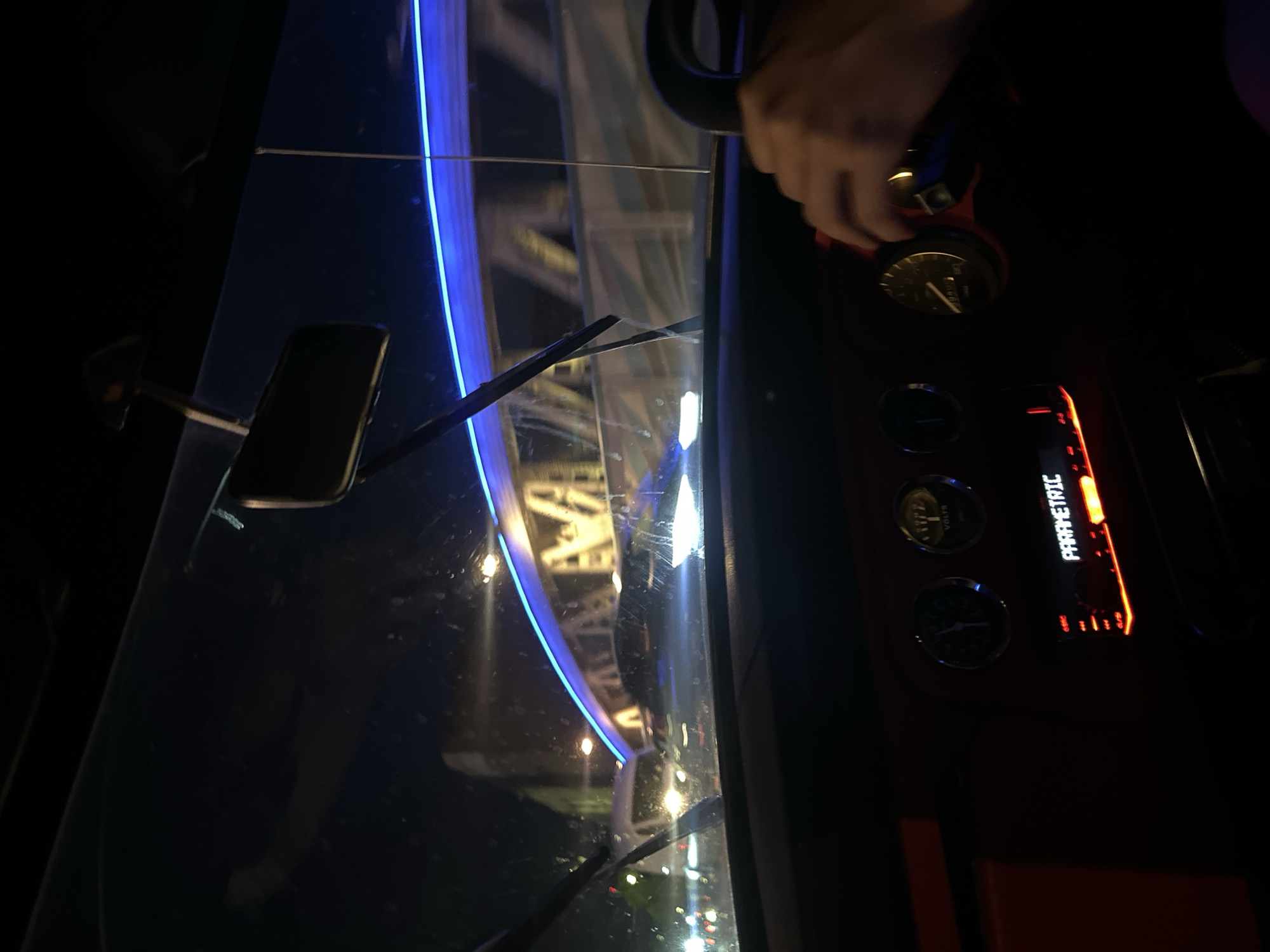 The image shows the interior of a vehicle at night, possibly a car, with a view through the windscreen of what appears to be the London Eye in London, United Kingdom, illuminated with blue lights. Inside the vehicle, a hand is visible manipulating a gauge on a red-trimmed dashboard, which also features other gauges and an illuminated display with the word "PARAMETRIC". The windscreen shows reflections of lights and structures, suggesting a wet or reflective surface. The general environment is dark, with artificial lights providing the main source of illumination.