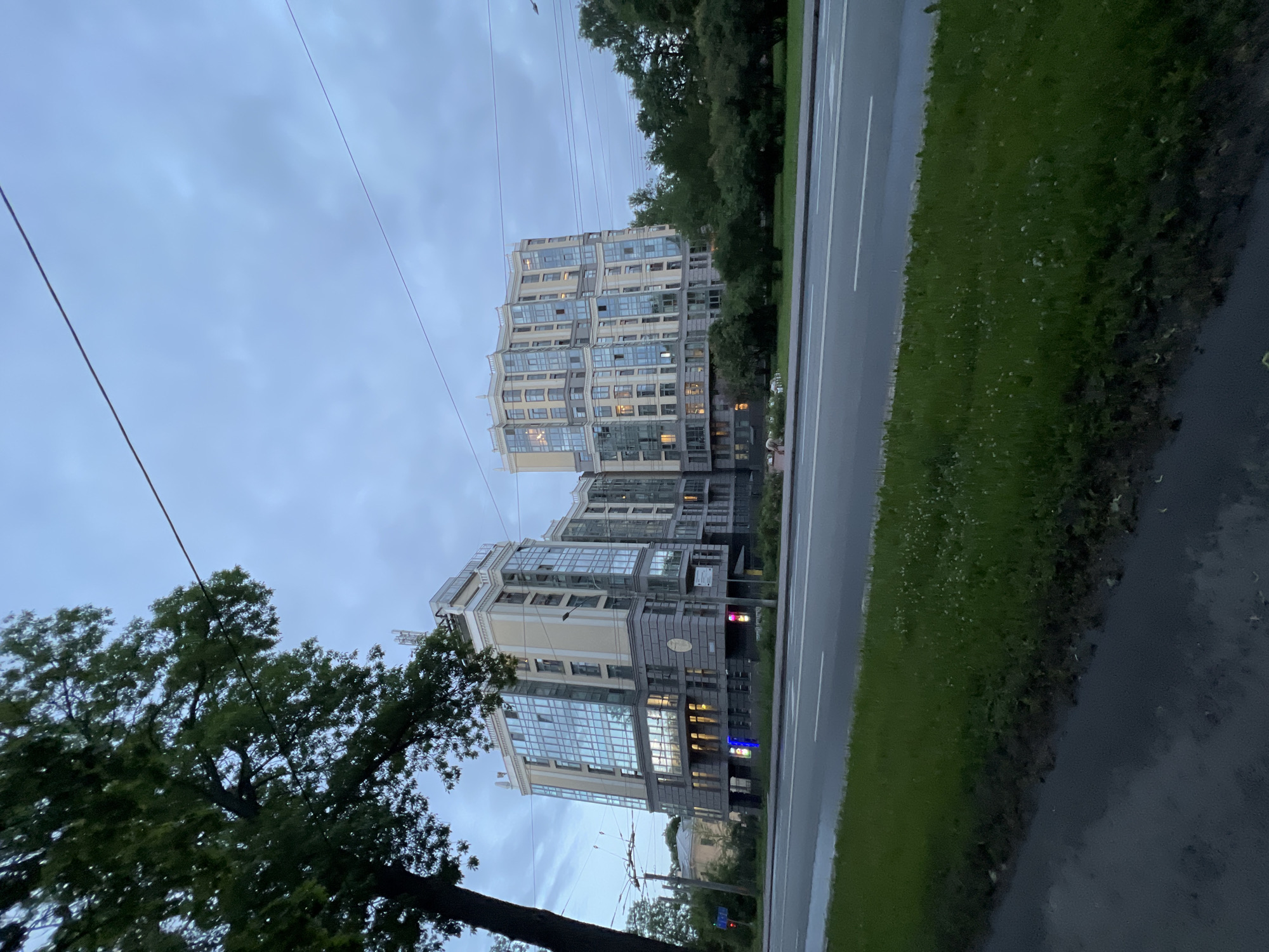 A modern, multi-story residential building complex with a beige facade and extensive glass windows is situated next to a wide road. The building has an undulating roofline and is topped with several antennas. A mature tree with dark green foliage partially obscures the left side of the building and extends into the sky. The sky is overcast and a light blue color, with thin clouds. Power lines are visible stretching across the sky, originating from poles near the tree on the left. The road in the foreground has a grey asphalt surface with white lane markings. A strip of green grass, dotted with small white flowers, separates the road from a darker, possibly wet, paved area on the far left. The scene appears to be set in a city, likely Saint Petersburg, Russia, given the architectural style and the presence of overhead power lines typical of the city. No people are clearly visible, though a small white object, possibly a pet or a person, is visible on the grass in front of the building on the right. The lighting suggests it is either early morning or late evening due to the diffused light and overcast conditions.